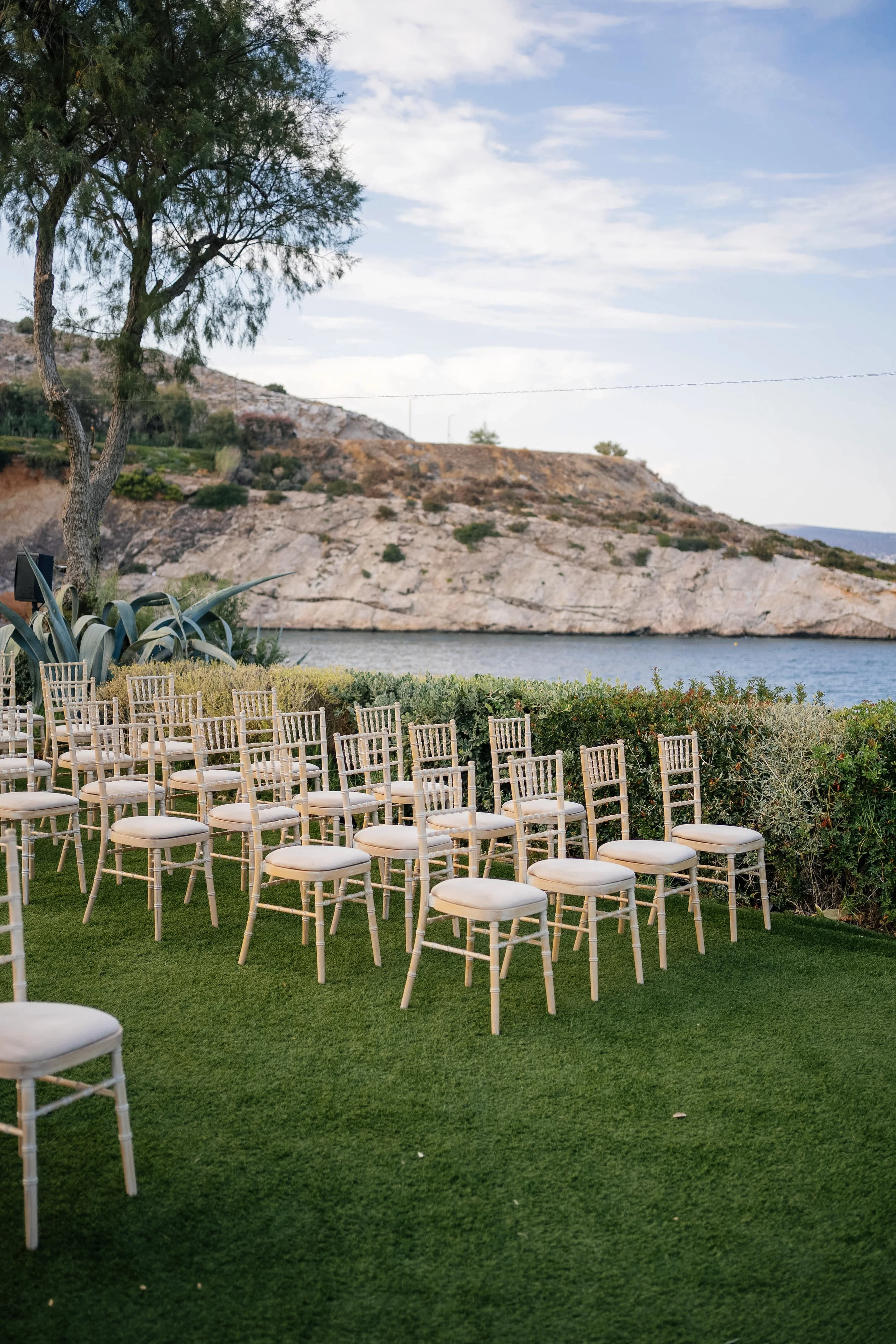 Empty white chairs set up outdoors on green grass, with a backdrop of water, rocky hills, trees, and a partly cloudy sky.