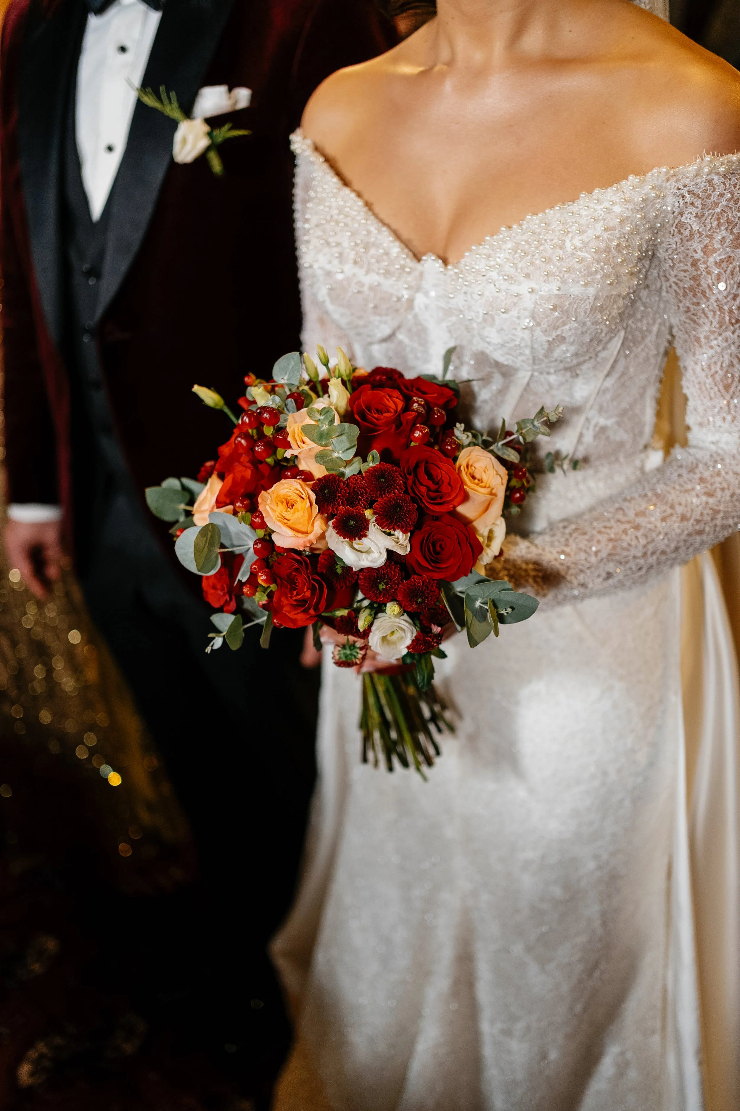 Close-up of a bride in a strapless, white wedding gown holding a bouquet of red, peach, and white roses, with greenery. The background includes a groom in a black tuxedo with a white shirt and boutonniere.