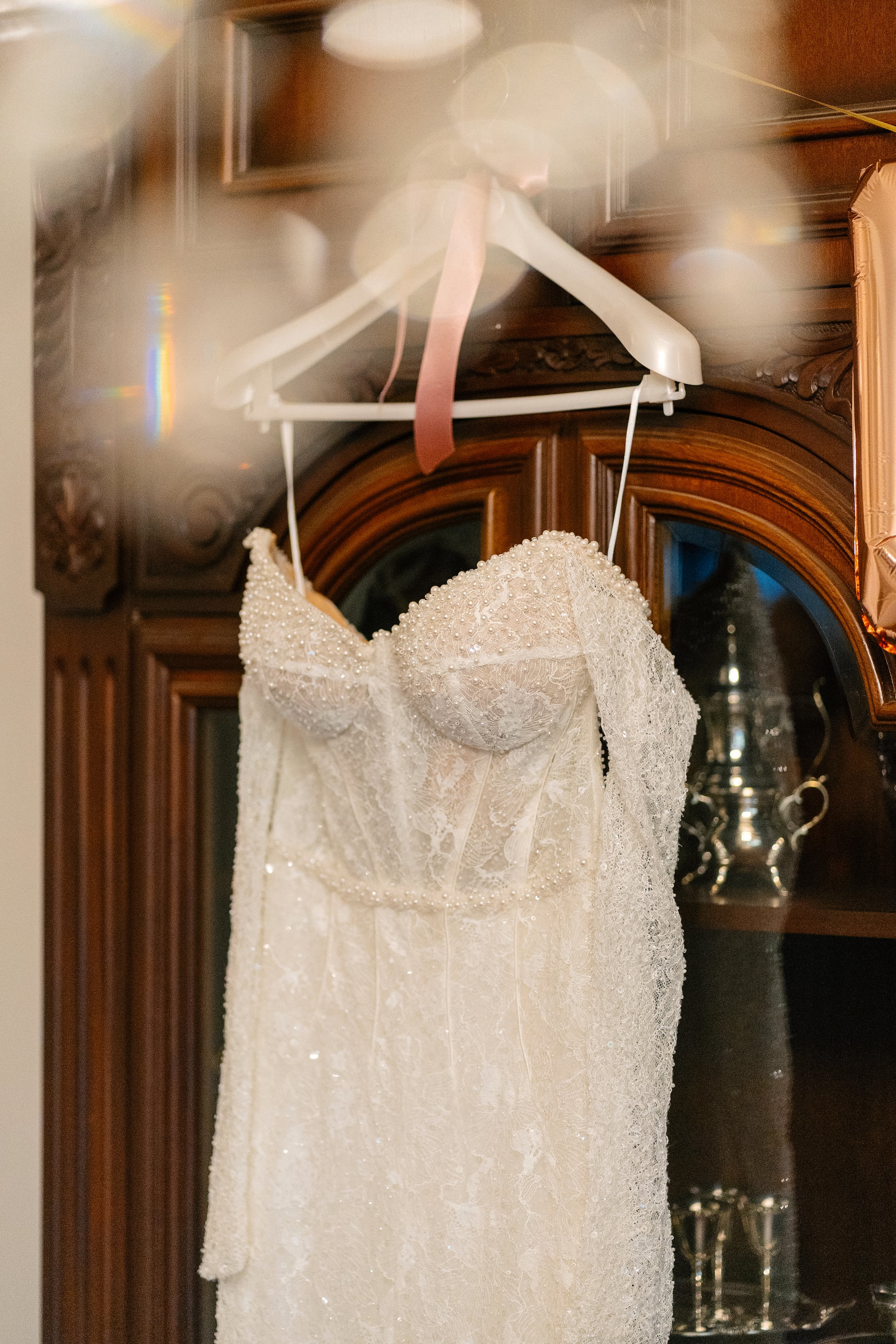 A wedding dress hanging on a white hanger, decorated with pearls and lace, displayed in front of a wooden cabinet with glass doors.