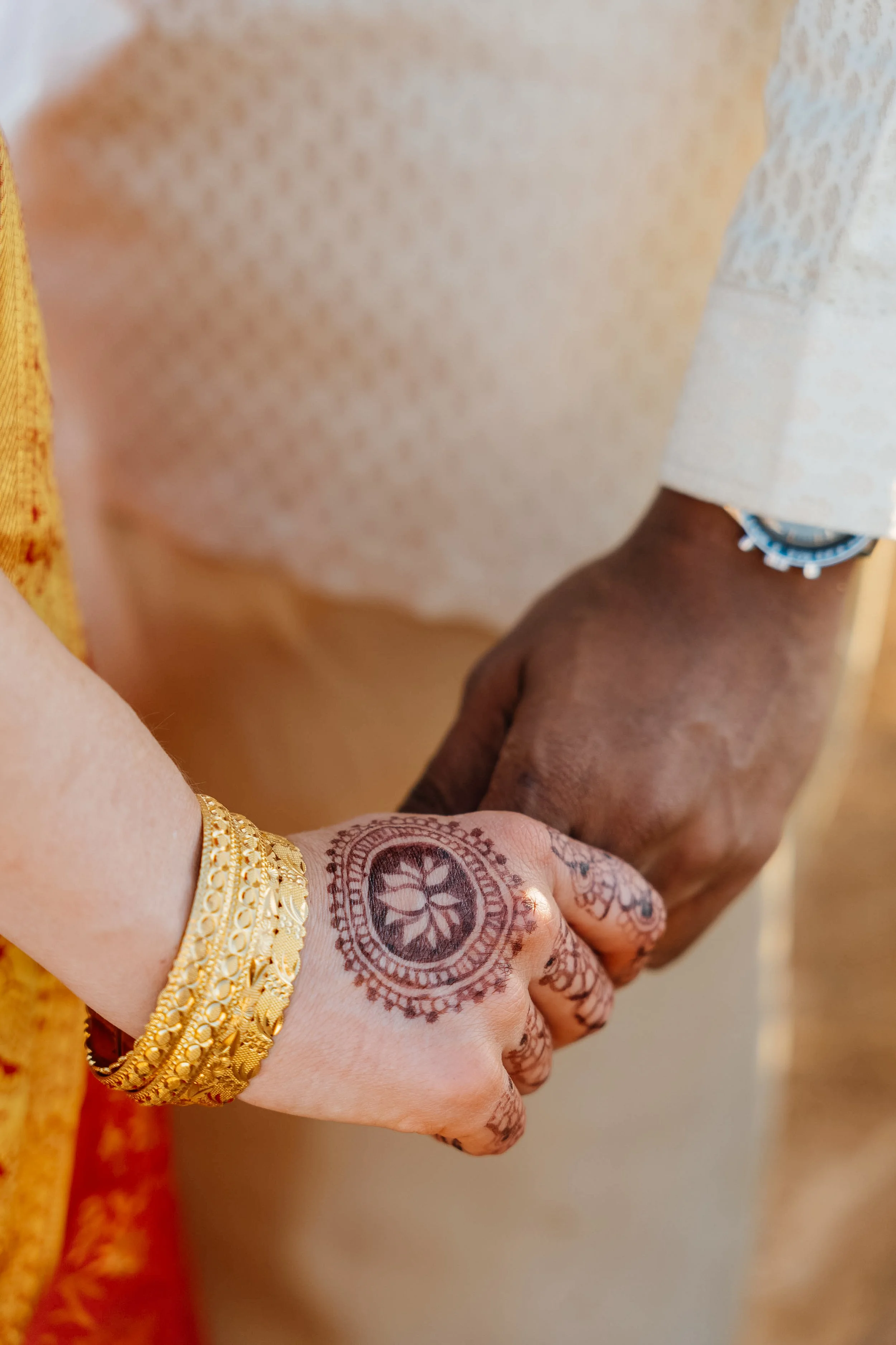 Close-up of two hands holding each other, one with henna designs and a gold bracelet, and the other with a darker skin tone, wearing a bracelet. The person with henna is wearing traditional clothing with gold embroidery.