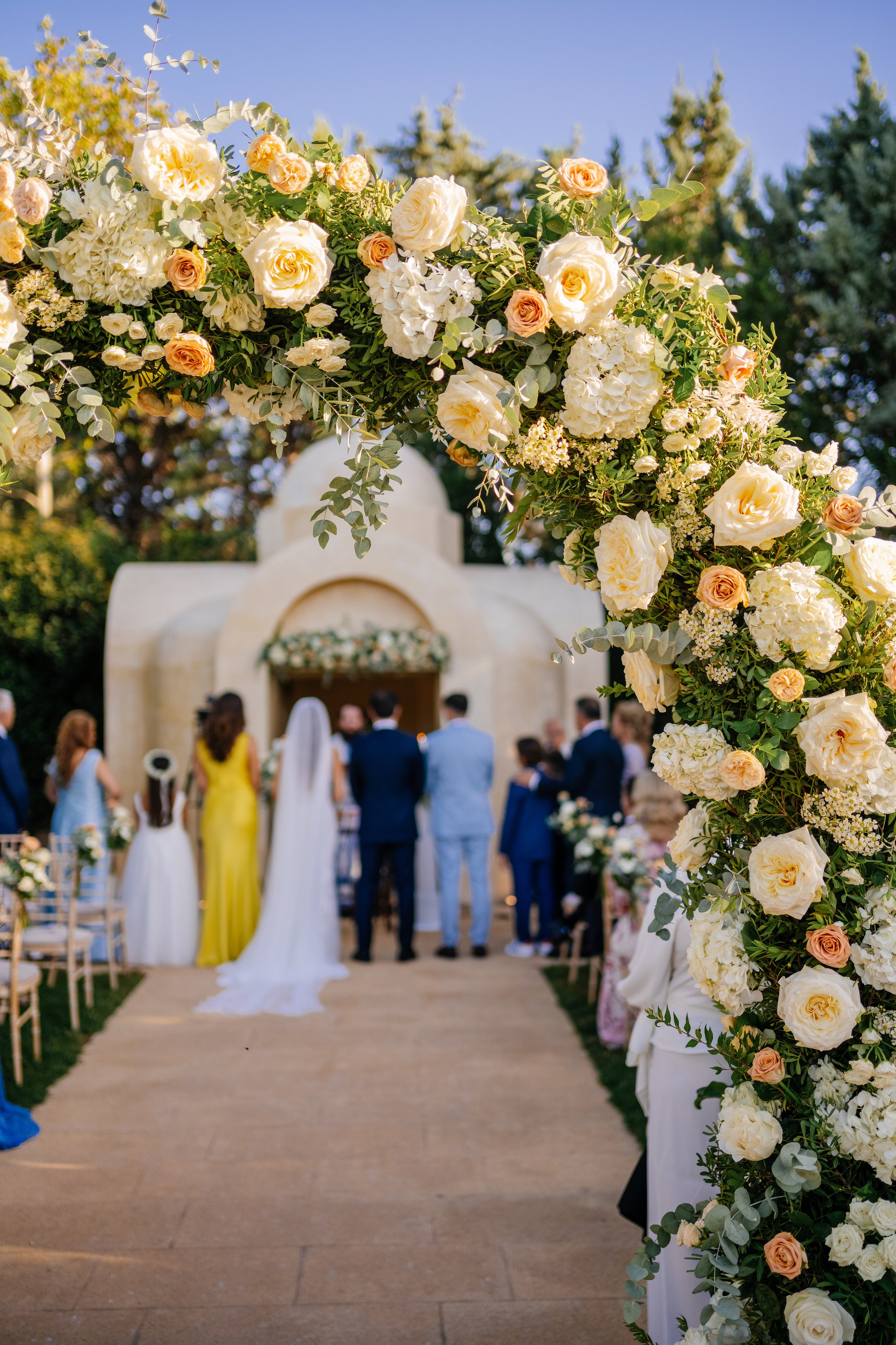 Wedding ceremony outdoor with an arch decorated with white and peach flowers, and guests standing in front of an altar.