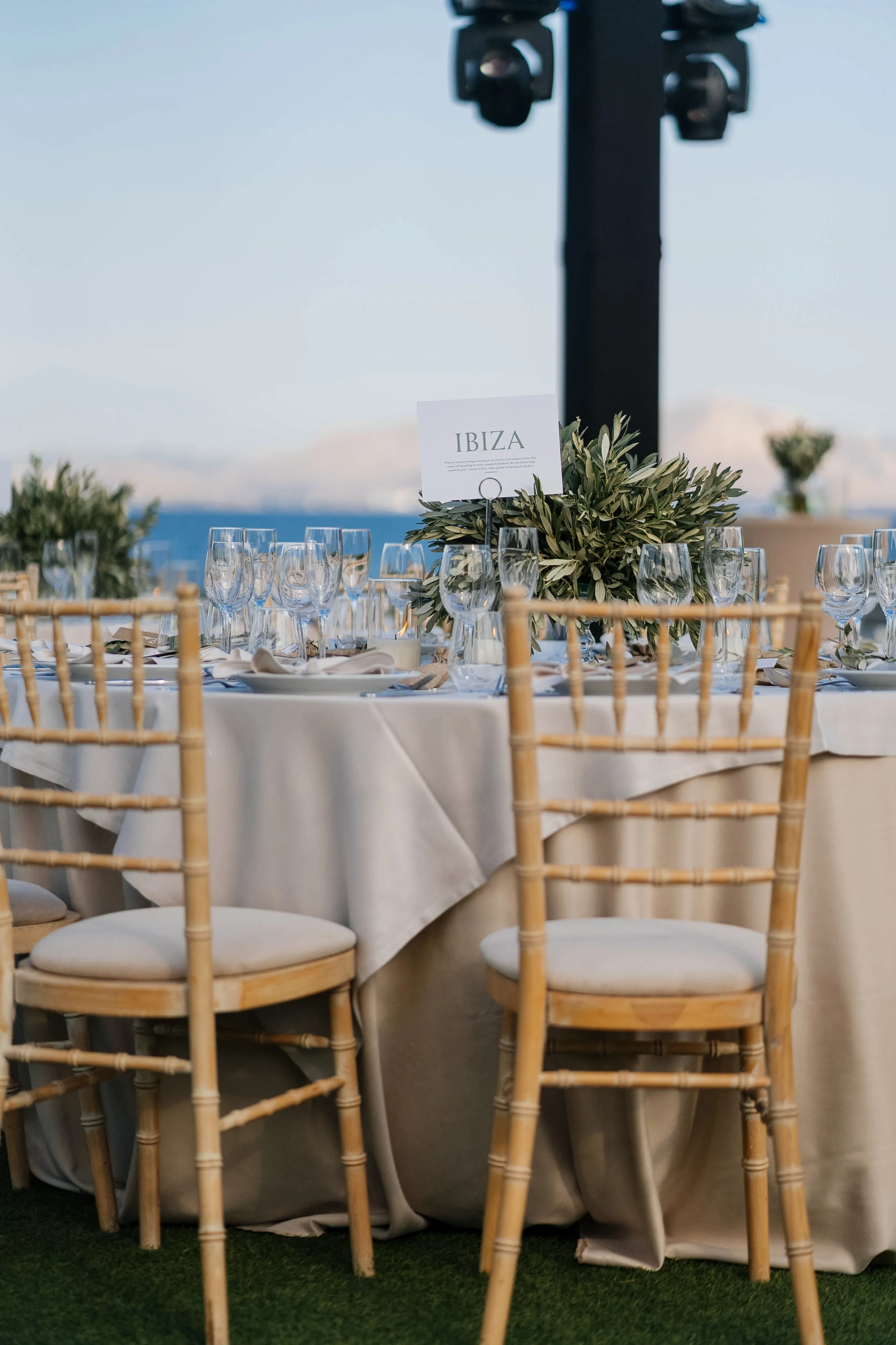 Wedding reception table with glassware, a sign labeled 'IBIZA,' greenery centerpiece, set against an outdoor ocean backdrop with sky and clouds.