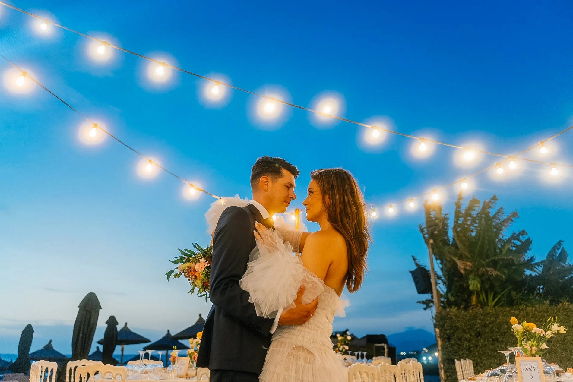 A couple dressed in wedding attire, standing close and gazing at each other during an outdoor wedding reception at dusk, with string lights overhead and tables with white chairs and floral centerpieces in the background.