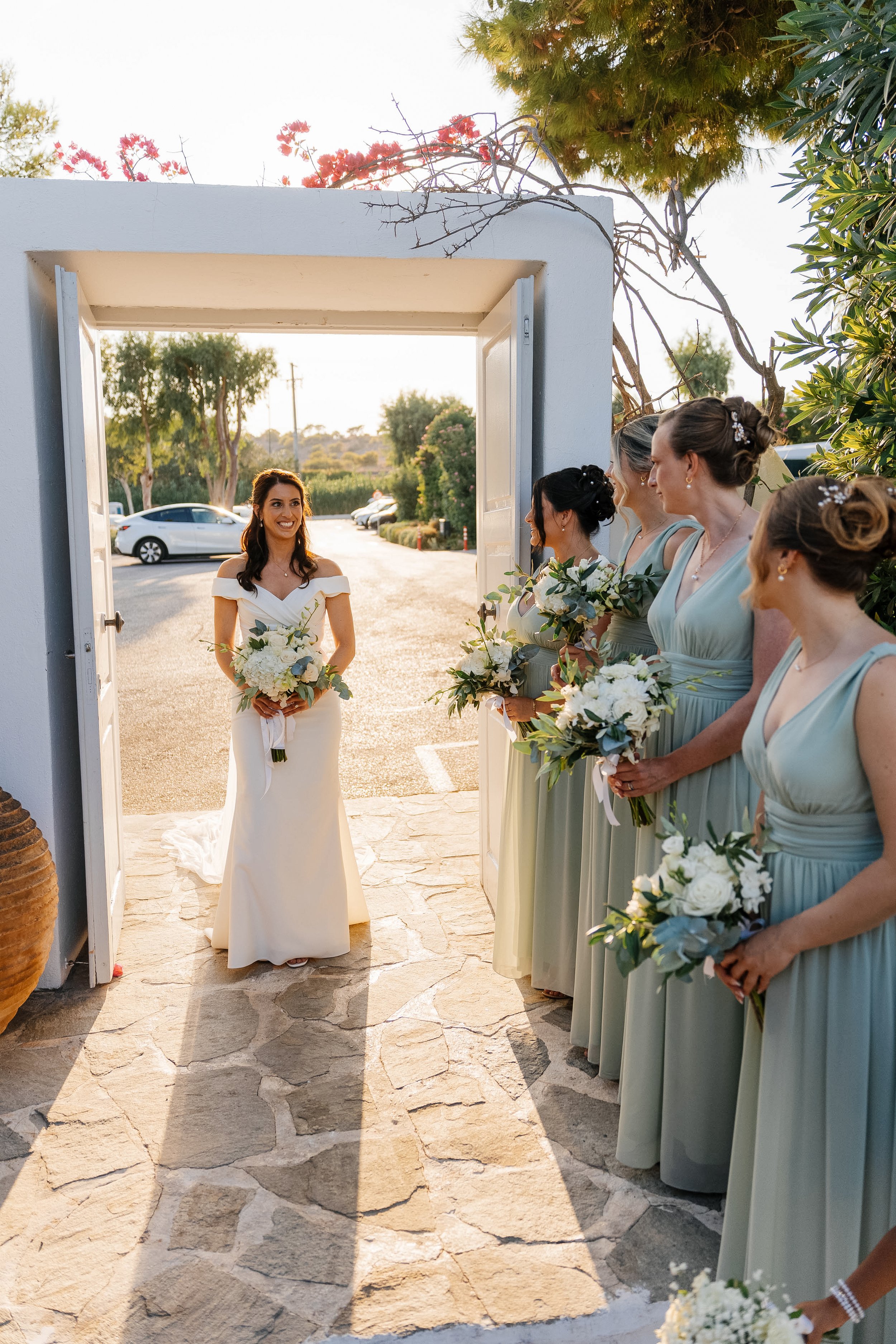 A bride standing at the entrance of a building, smiling, holding a bouquet, with five bridesmaids dressed in light blue gowns holding flowers, standing on the sidewalk outside during sunset. Trees and parked cars are visible in the background.