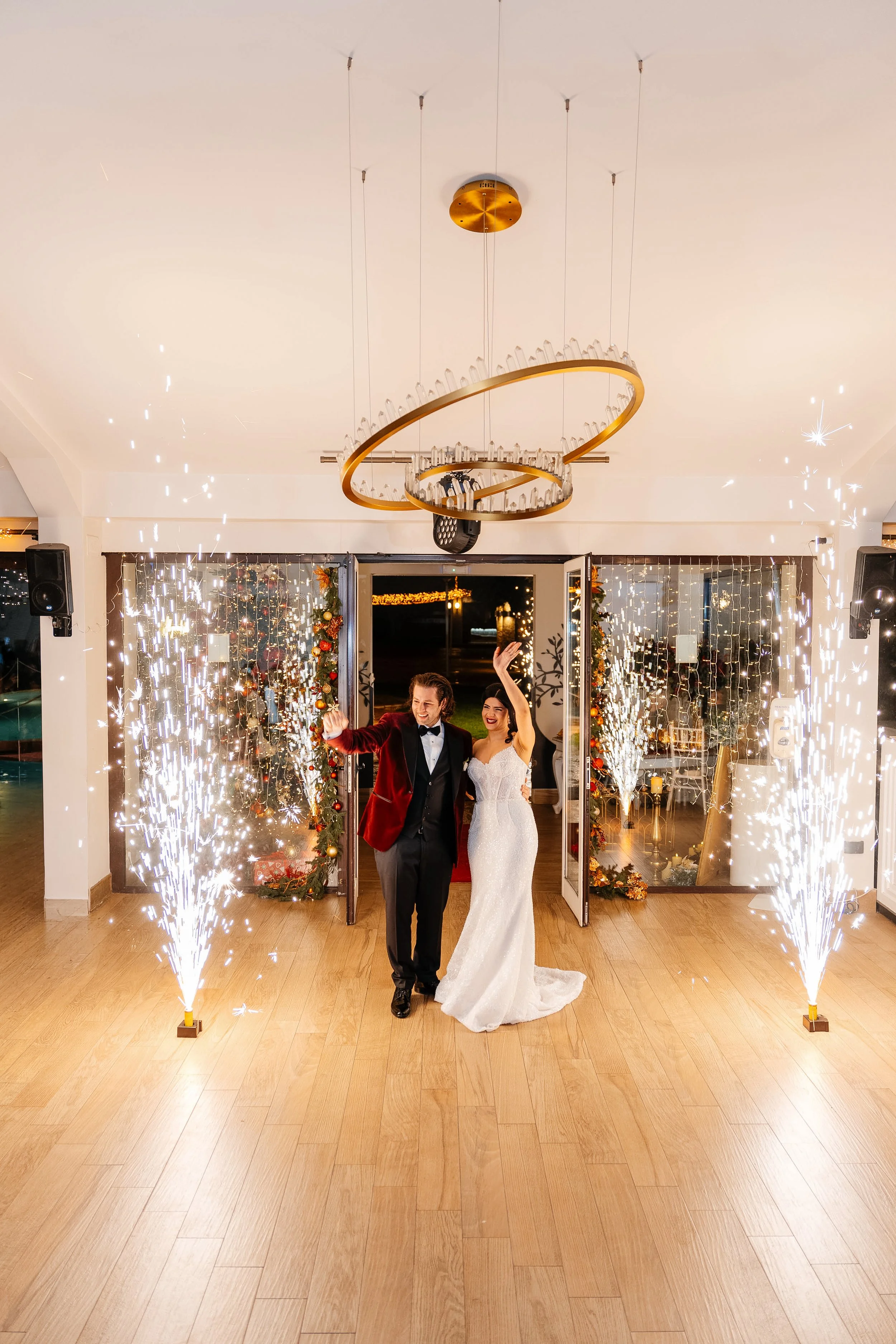 Happy bride and groom walking through a celebration with fireworks and sparklers, decorated with Christmas ornaments, at a wedding venue.