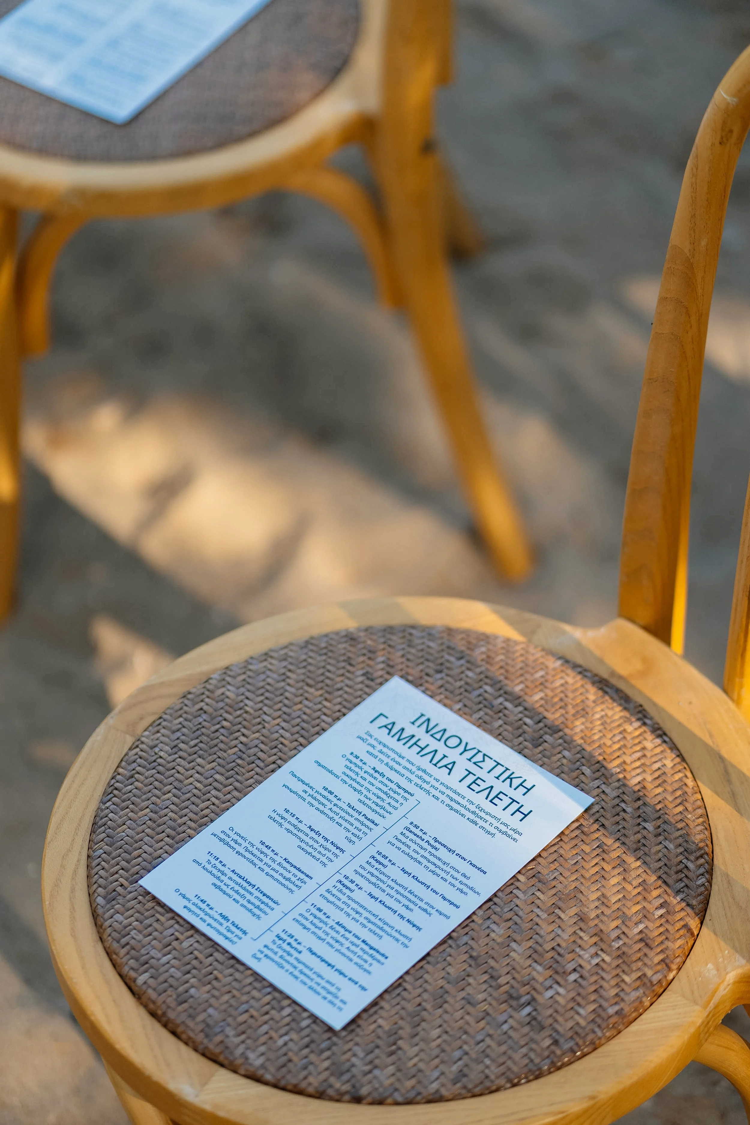 A wooden chair with a woven seat holds a piece of paper with Greek text. Another similar chair is partly visible in the background.