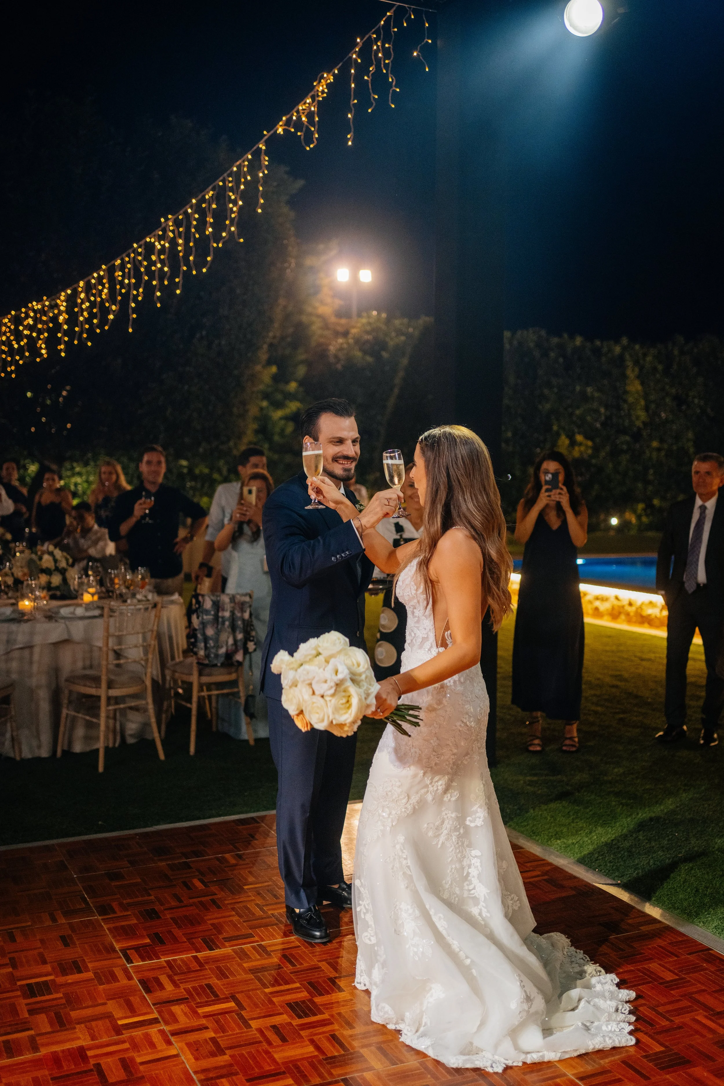 A bride and groom sharing a toast with champagne at their outdoor wedding reception during nighttime, surrounded by guests and string lights.