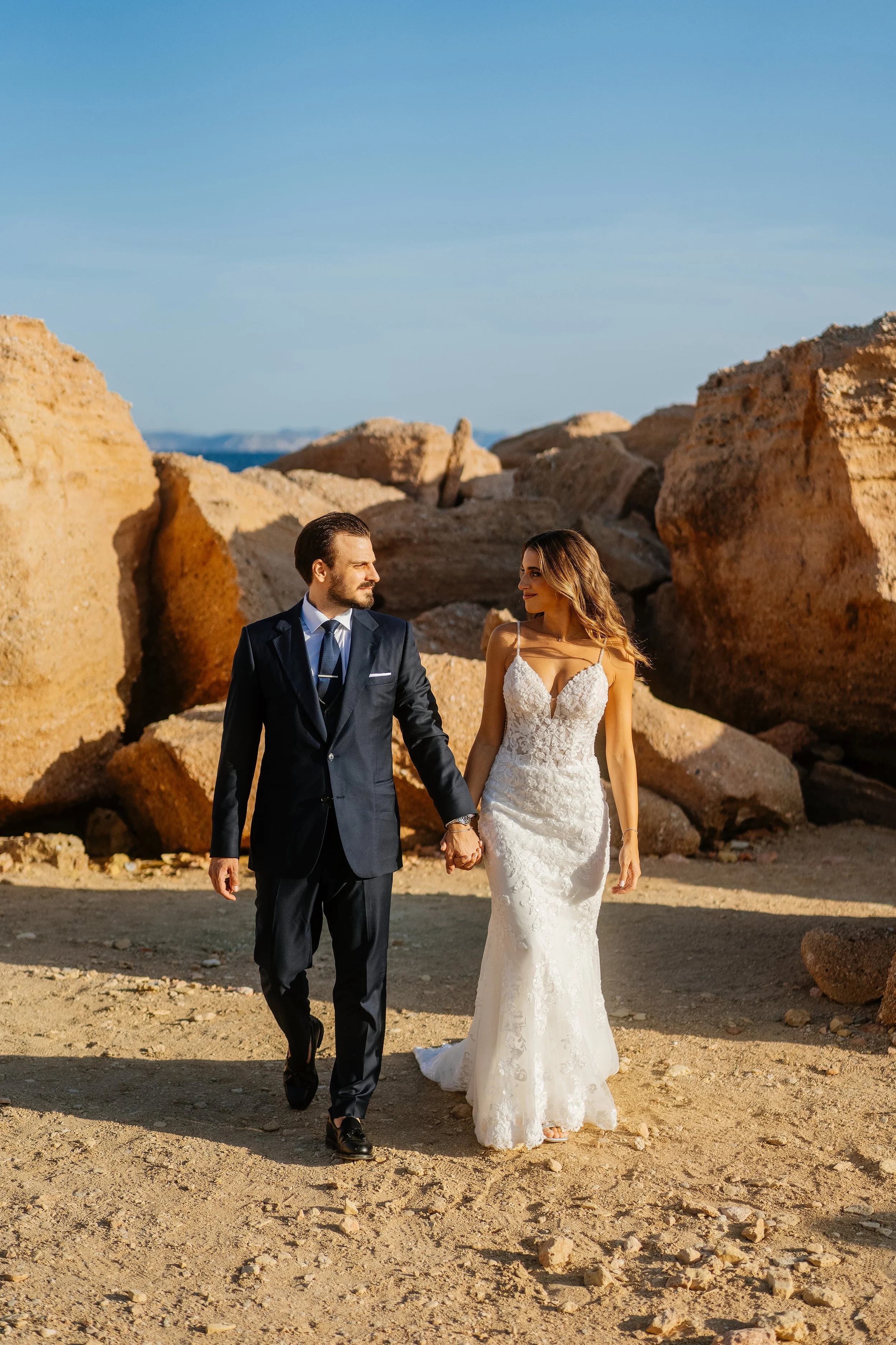 A couple in wedding attire walking hand in hand outdoors in a rocky desert landscape under a clear blue sky.
