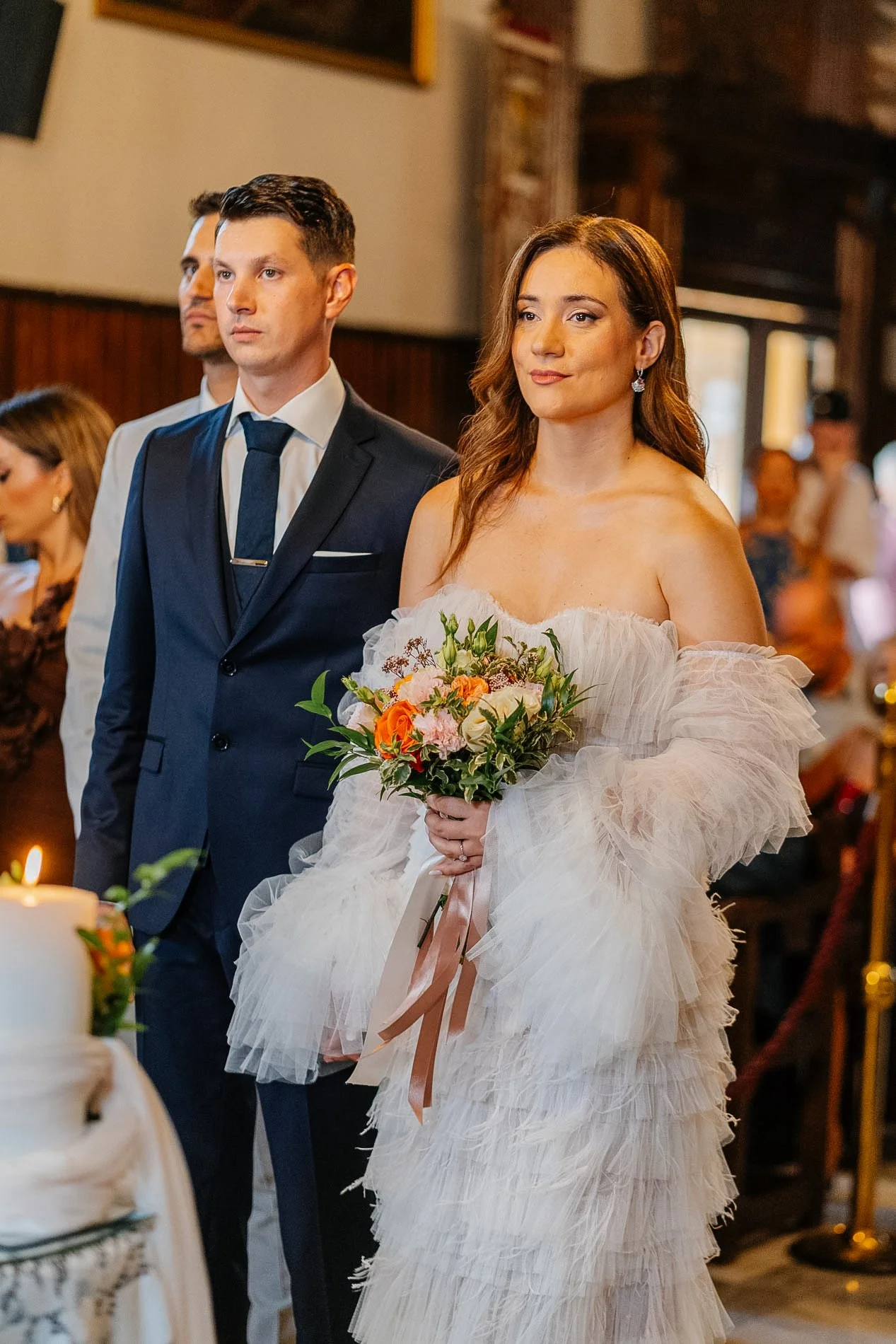 A bride holding a bouquet of flowers in a wedding ceremony indoors, standing in front of groom and guests, with candles and decor in the background.