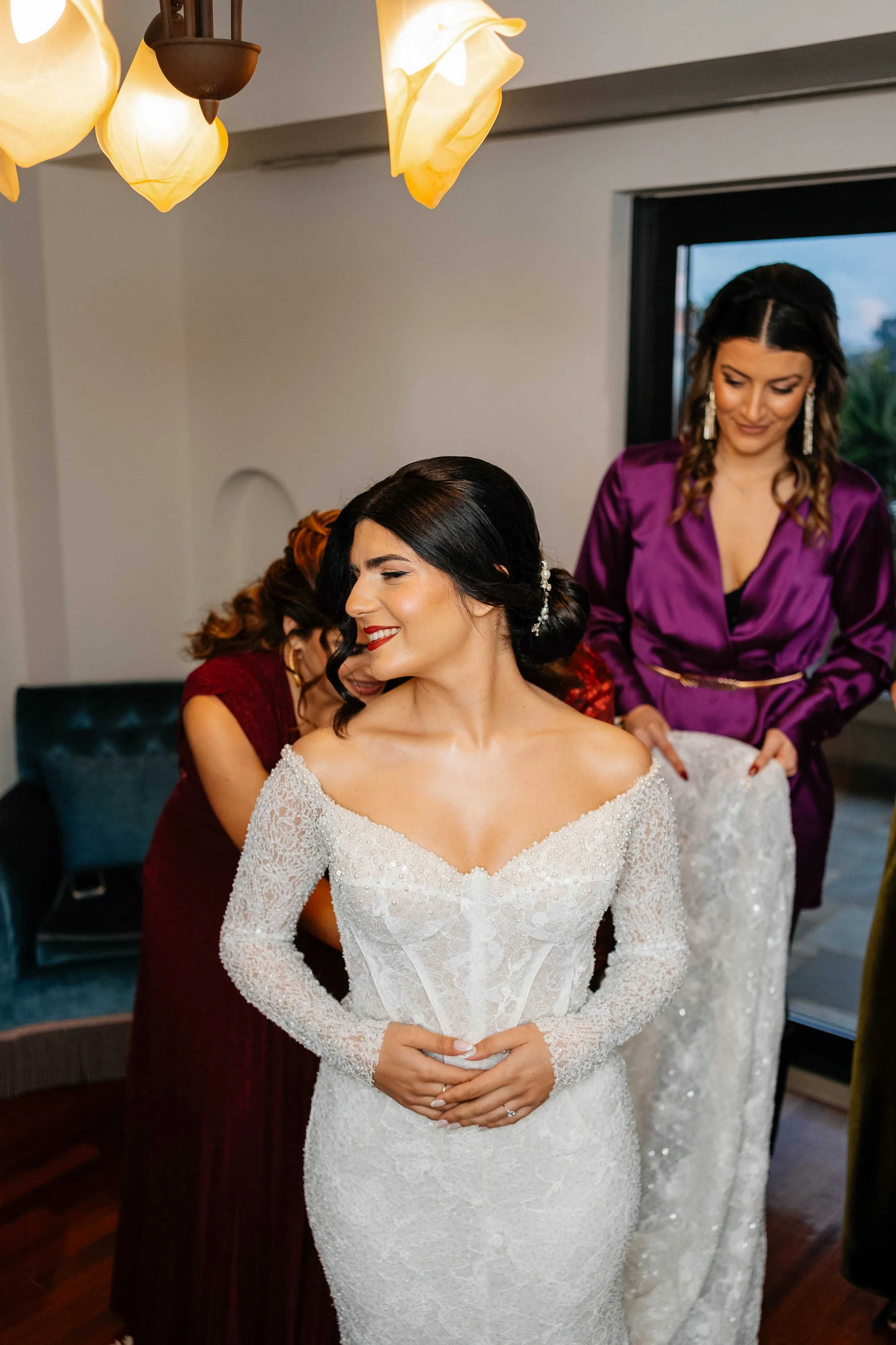 A bride in a lace wedding gown with long sleeves, standing with her hands clasped in front of her. A woman in a purple satin dress is helping her with her gown. Other women are partially visible in the background.