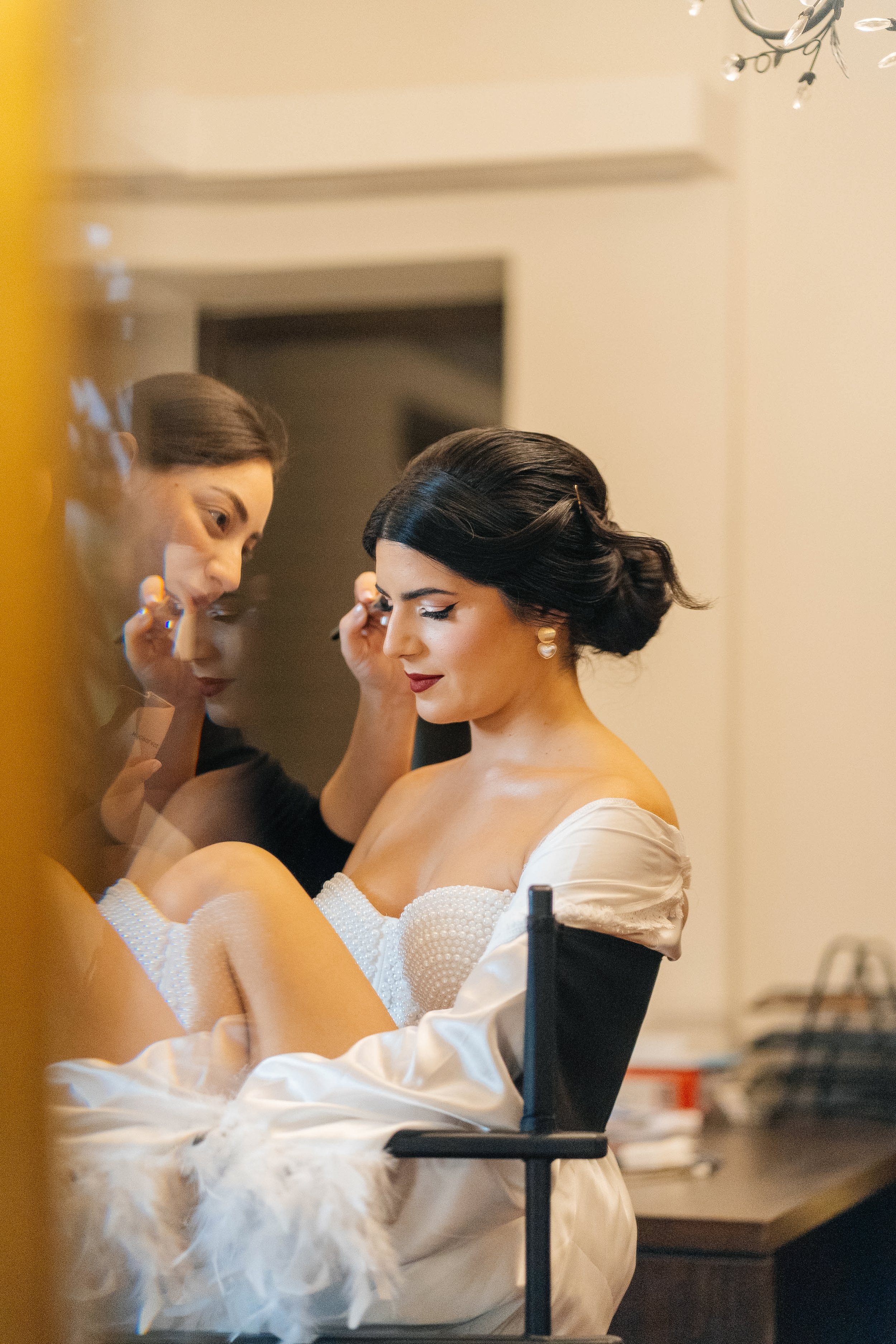 A woman is getting her makeup done by another woman in an elegant room. The woman receiving makeup is wearing a white dress with an off-the-shoulder design and pearl embellishments, and has dark, styled hair. The makeup artist is focused on her work,