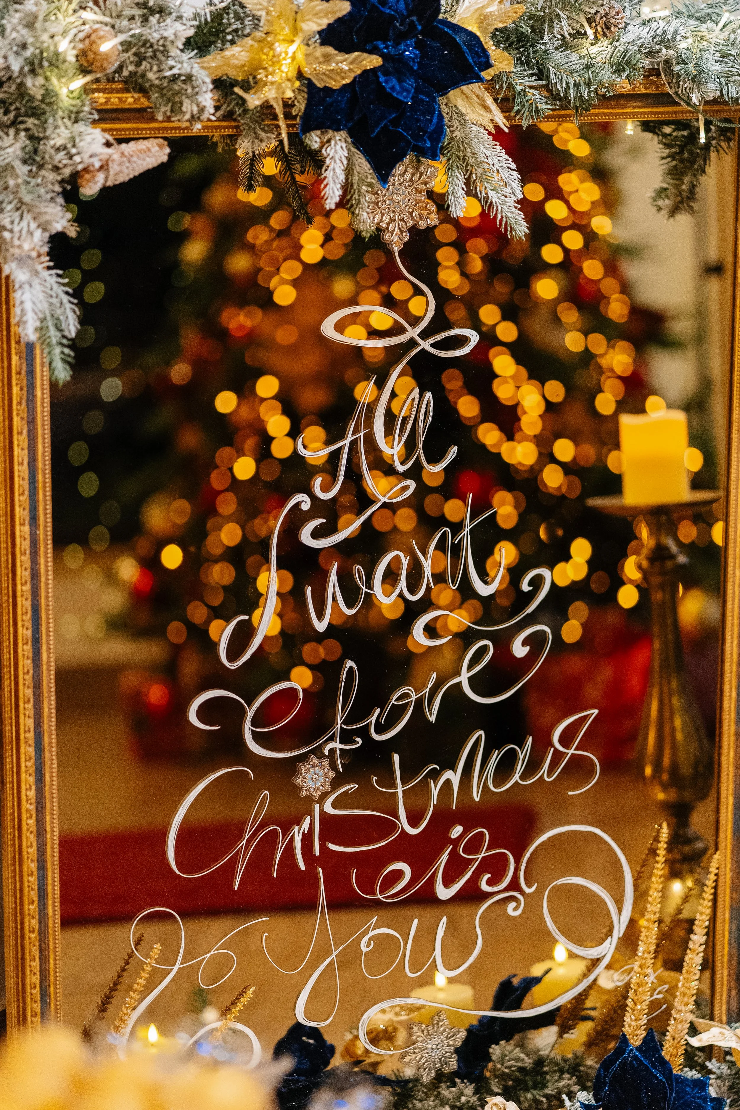 Holiday decorated mirror with snow-dusted greenery, gold and blue ornaments, and the phrase 'All I want for Christmas is You' written on it in cursive. In the background, blurred Christmas tree lights and a yellow candle are visible.