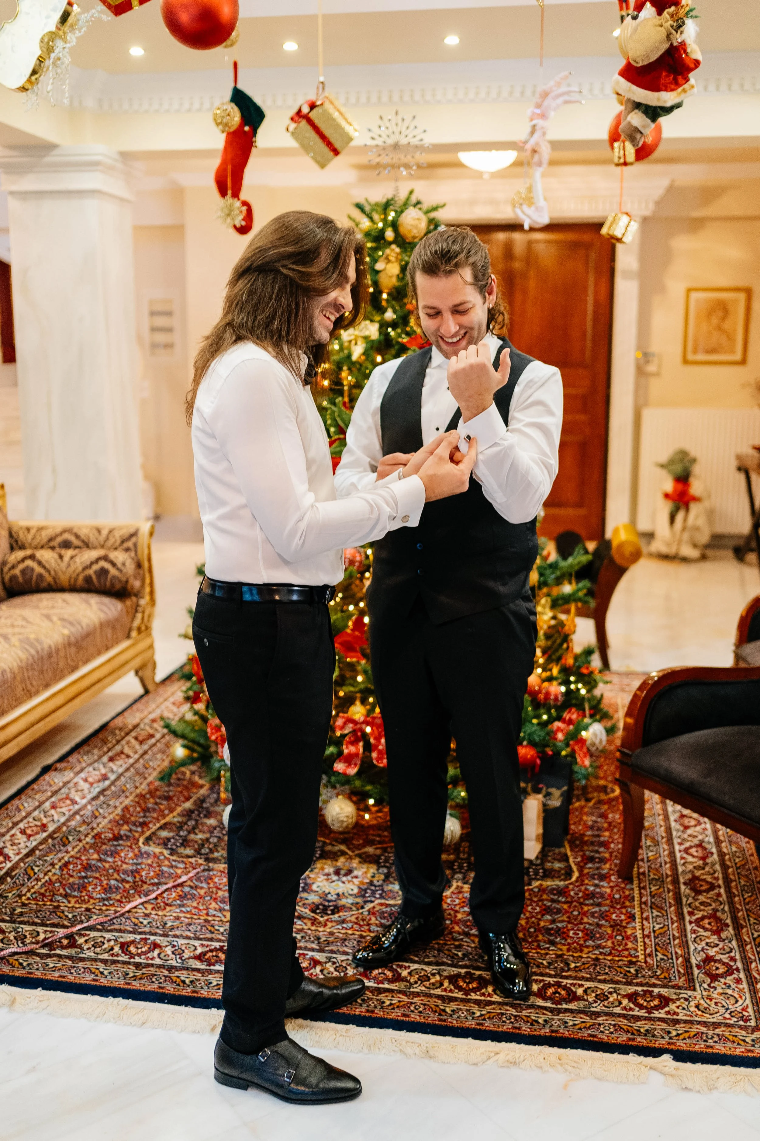 Two men in formal attire exchanging jewelry in a decorated Christmas living room with a Christmas tree and hanging ornaments.