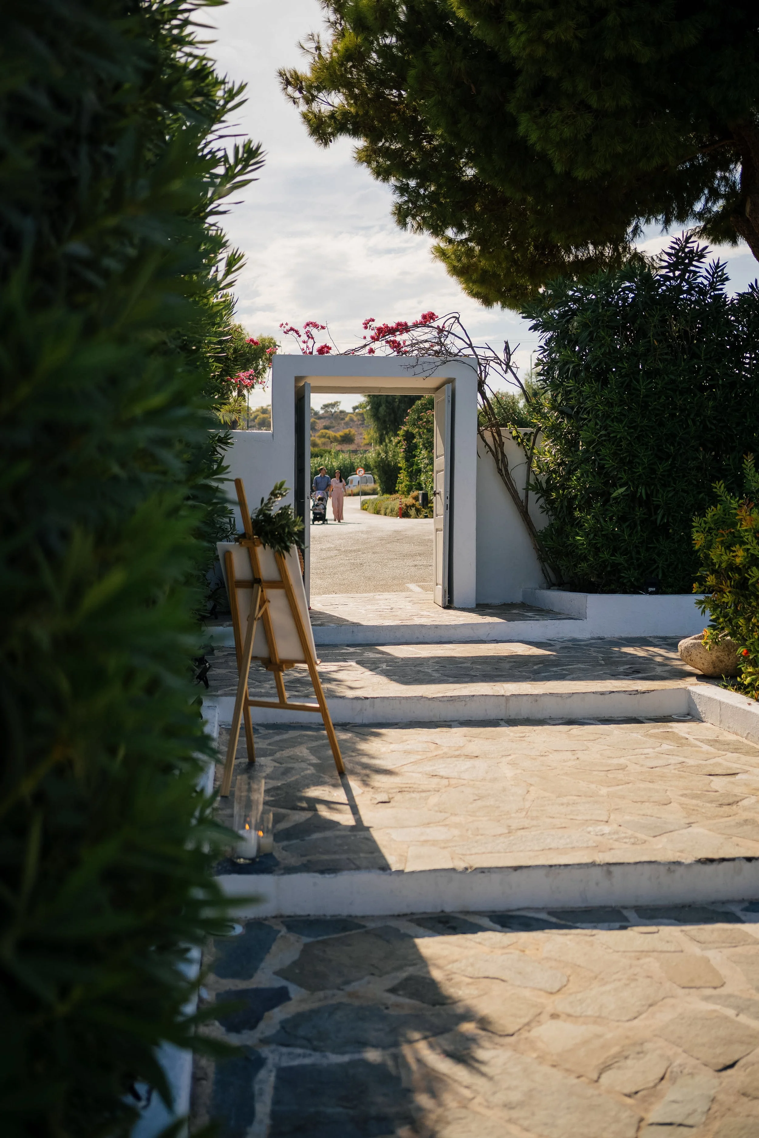 A view through an open white gate leading onto a stone pathway with greenery on both sides, two people walking in the distance, and a cloudy sky overhead.