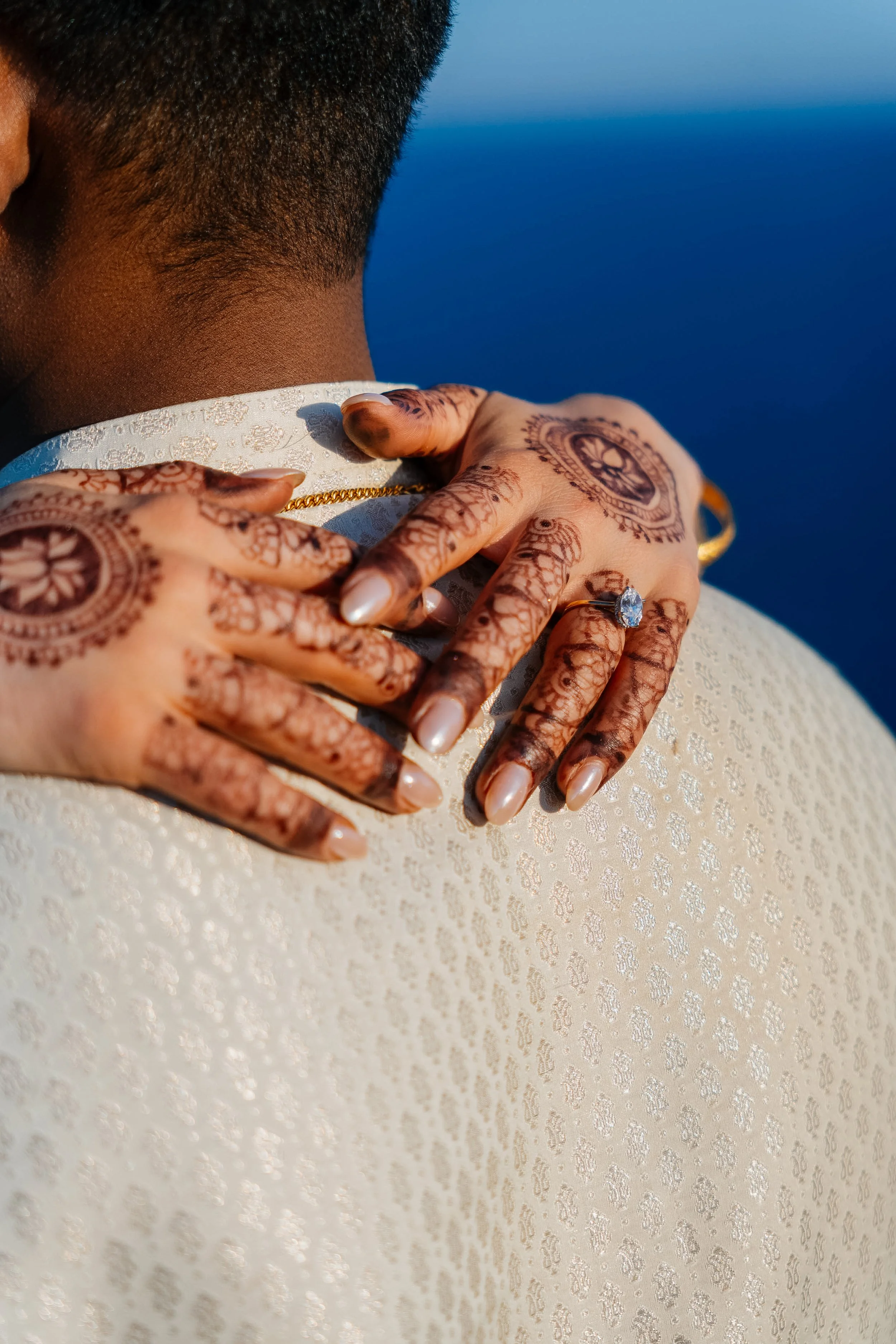 Close-up of a person's head and hands adorned with intricate henna designs, a ring with a large stone, and a gold chain, resting on textured white fabric against a blue background.