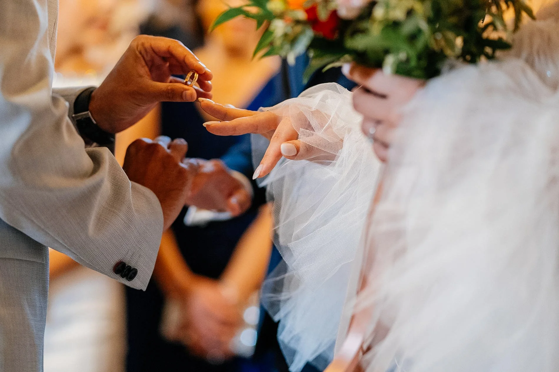 A wedding ceremony with a close-up of a couple exchanging rings. The groom is placing a ring on the bride's finger, and the bride is holding a bouquet of flowers. The bride's hand is adorned with a lace sleeve.