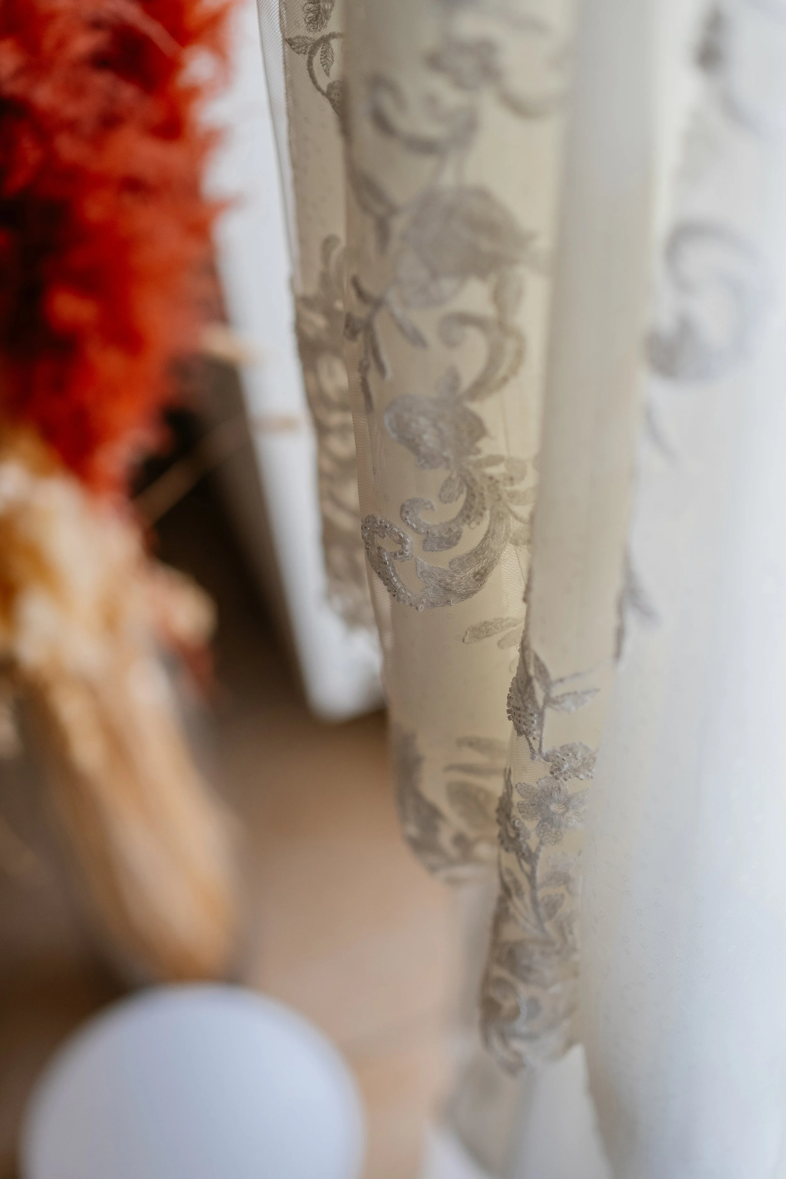 Close-up of a white lace curtain with floral embroidery, partially obscuring the view of some colorful objects on the left side, and a blurred sliver of a white object in the foreground.