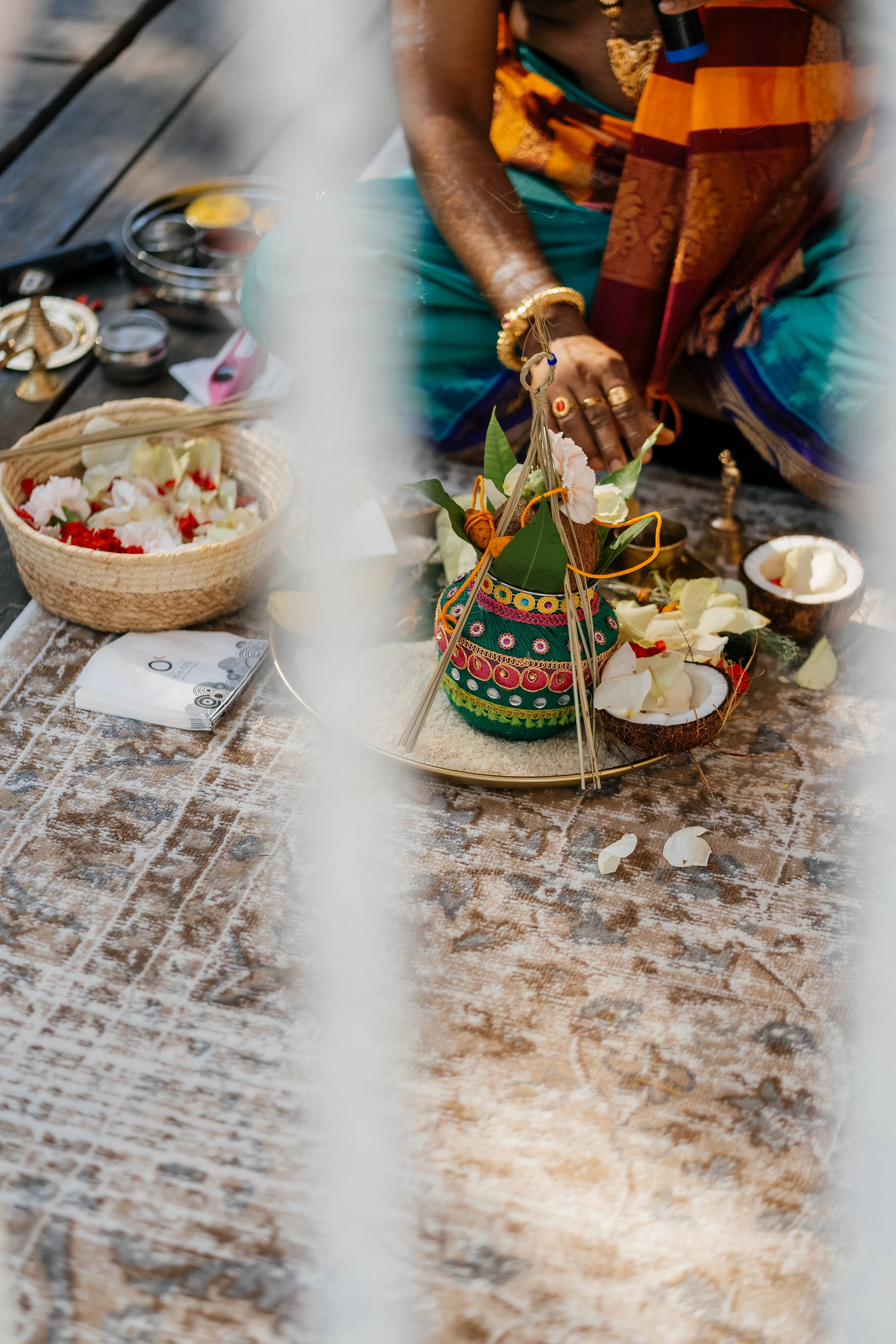 A person dressed in traditional, colorful clothing is performing a ritual or ceremony outdoors, with various ritual items and flower petals placed on a decorated cloth table.