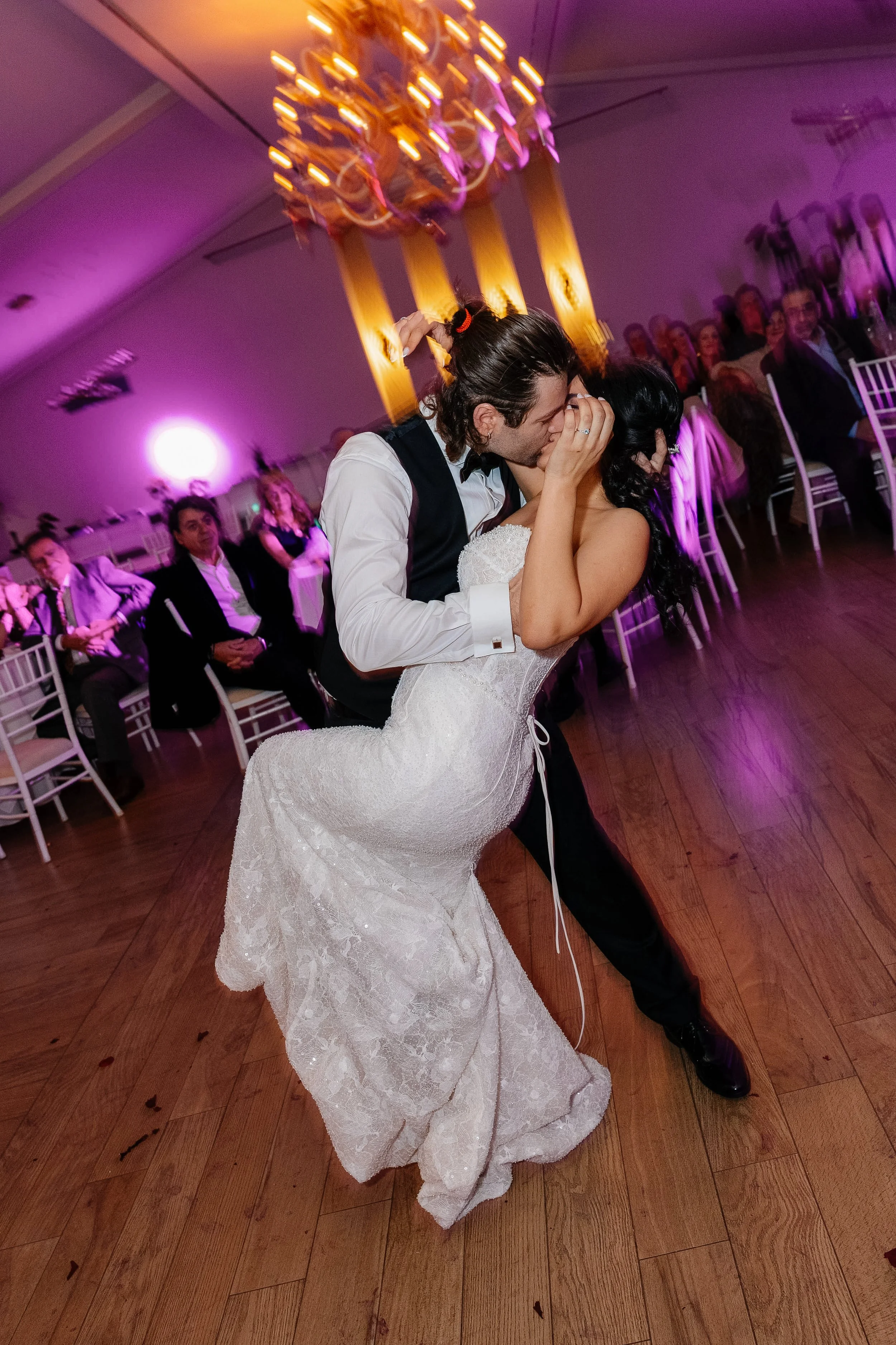 A newlywed couple sharing a dance at their wedding reception, with wedding guests seated at tables in the background and purple lighting creating a romantic atmosphere.