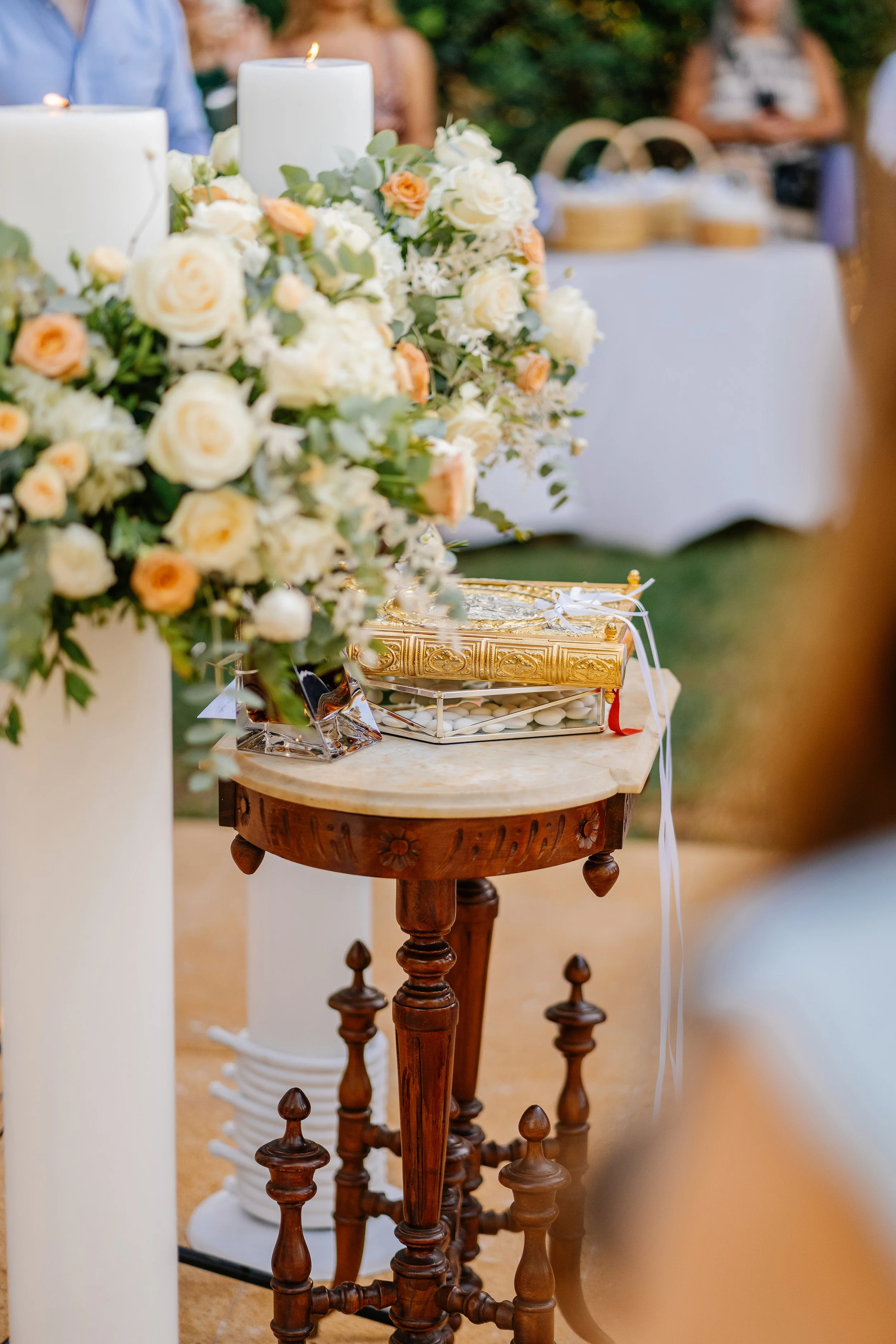 A table decorated with white and peach flowers, two white candles, and a box of wedding favors, set outdoors with people in the background.