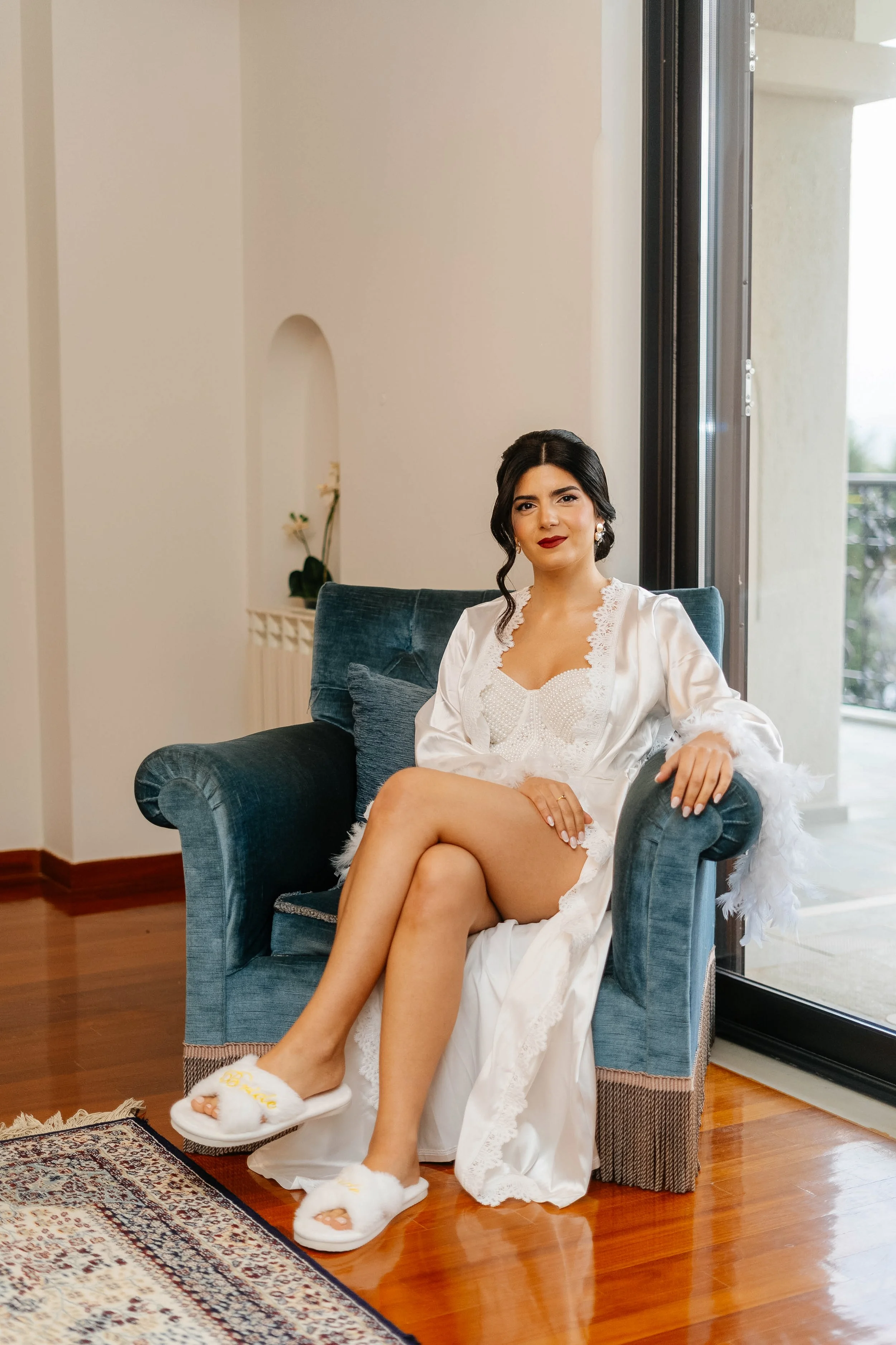 A woman with dark hair tied in a low bun, wearing pearls and red lipstick, sitting on a dark blue armchair in a bright room near a large window. She is dressed in white satin lingerie with lace details and fluffy white slippers. She has crossed her l
