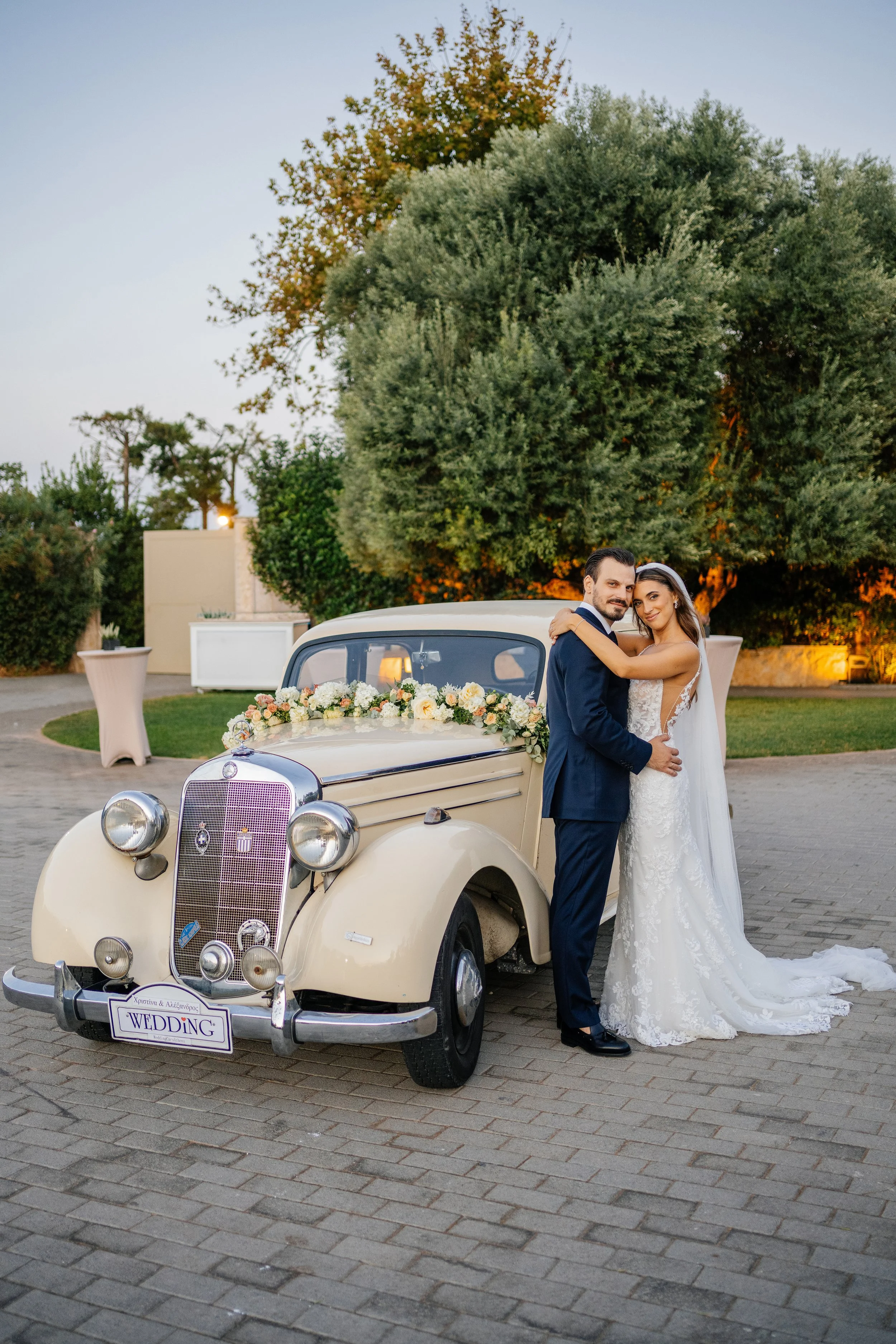 Bride and groom standing in front of a vintage cream-colored car decorated with flowers. The bride wears a white lace wedding gown and the groom a dark suit. They are embracing and smiling with trees and outdoor wedding decorations in the background.