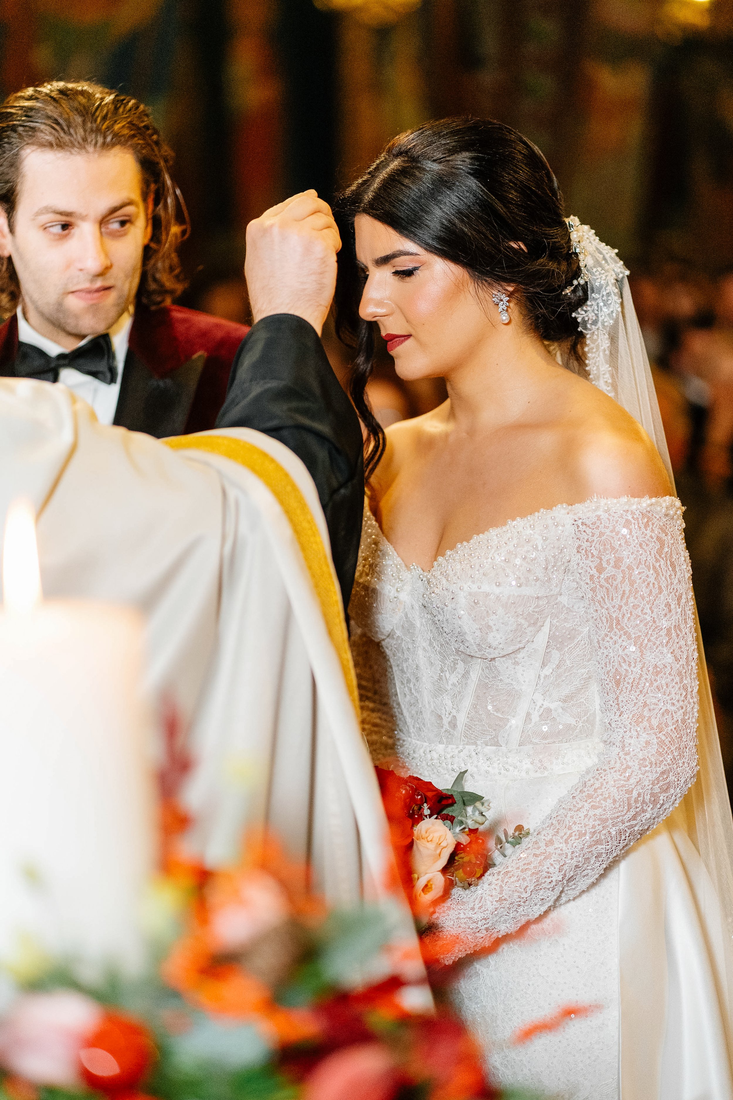 Bride and groom during wedding ceremony, bride wearing white lace wedding dress with off-the-shoulder sleeves, the groom in a tuxedo with a bow tie, officiant with white and gold robes, floral bouquet in the foreground.