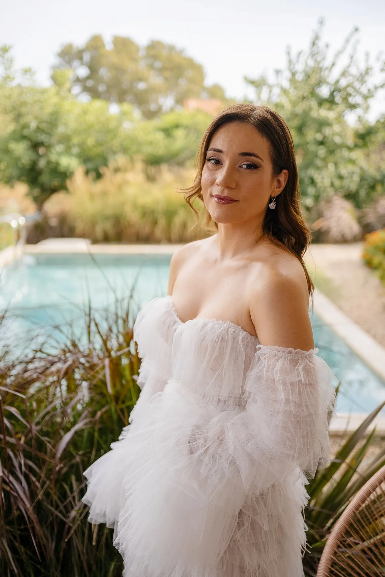 A woman with brown hair and earrings, wearing a white off-shoulder dress, standing outdoors near a pool with green trees and bushes in the background.