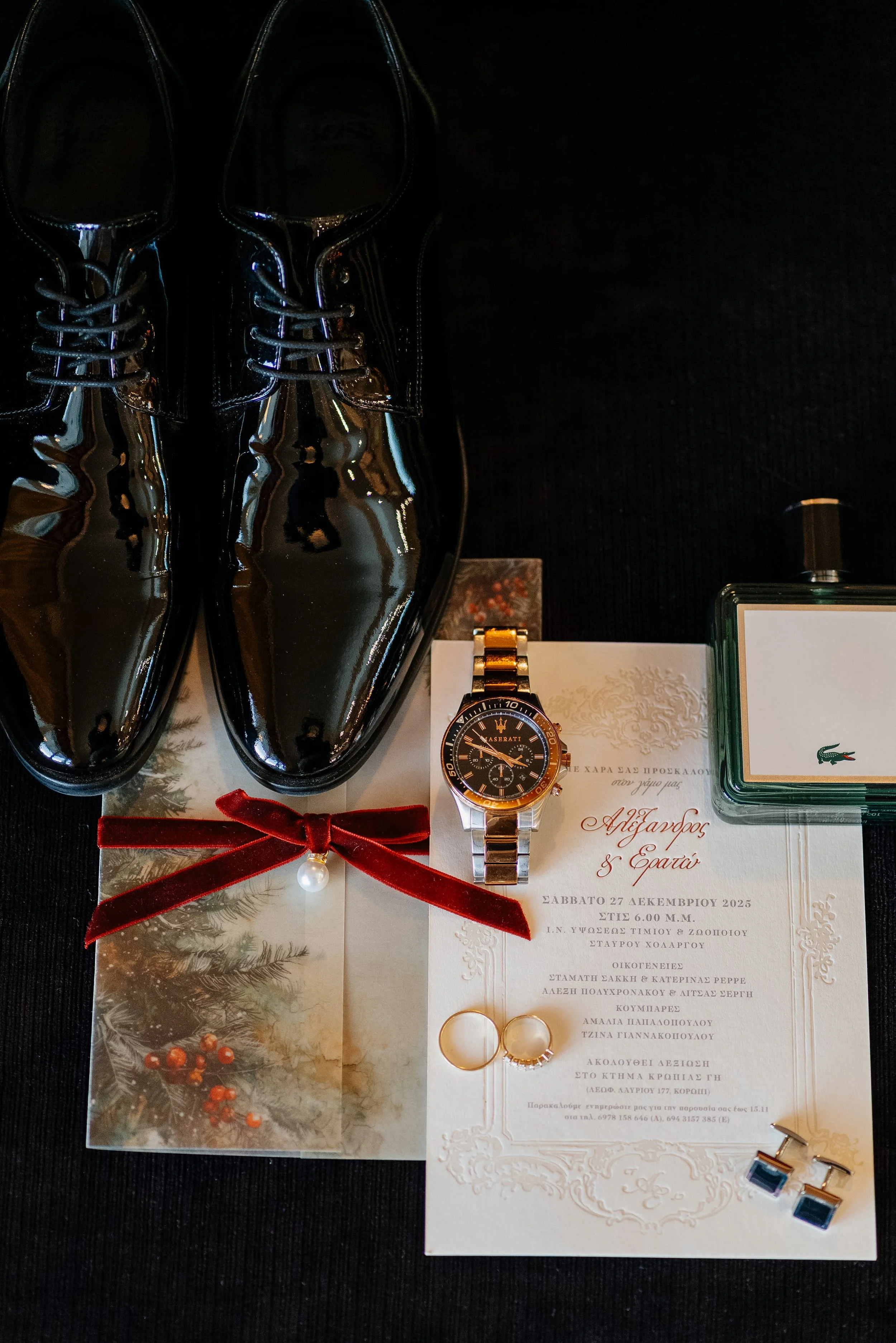 Pair of shiny black dress shoes, a sports watch with the Maserati logo, a wedding invitation, a pair of earrings with pearls, a ring, a celebratory card with a red ribbon, and Christmas-themed decorations on a black surface.