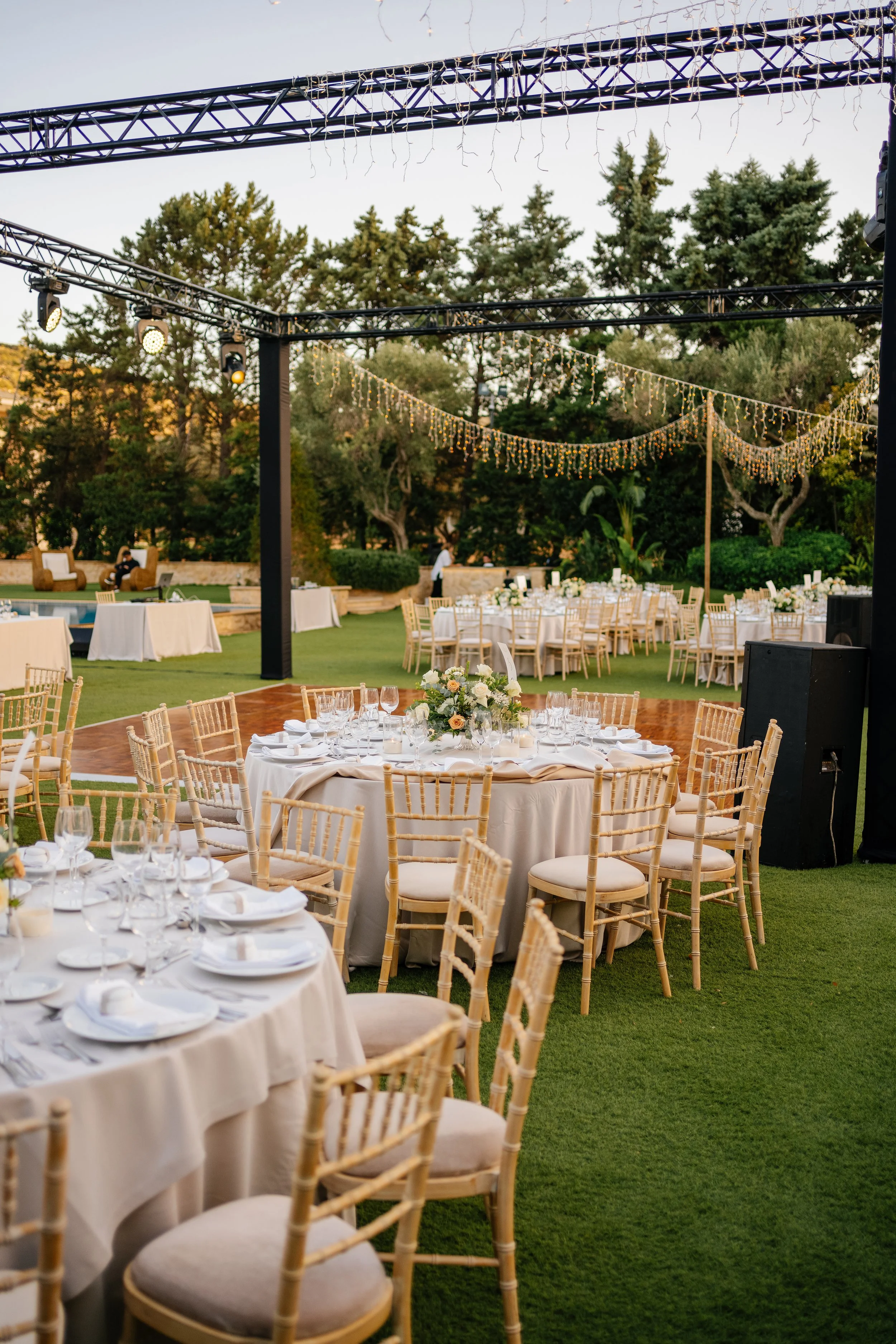 Outdoor event setup with round tables covered in white tablecloths, decorated with floral centerpieces, and surrounded by beige chairs with cushions. String lights hang above on black metal trusses, and a few people are seen in the background. The se