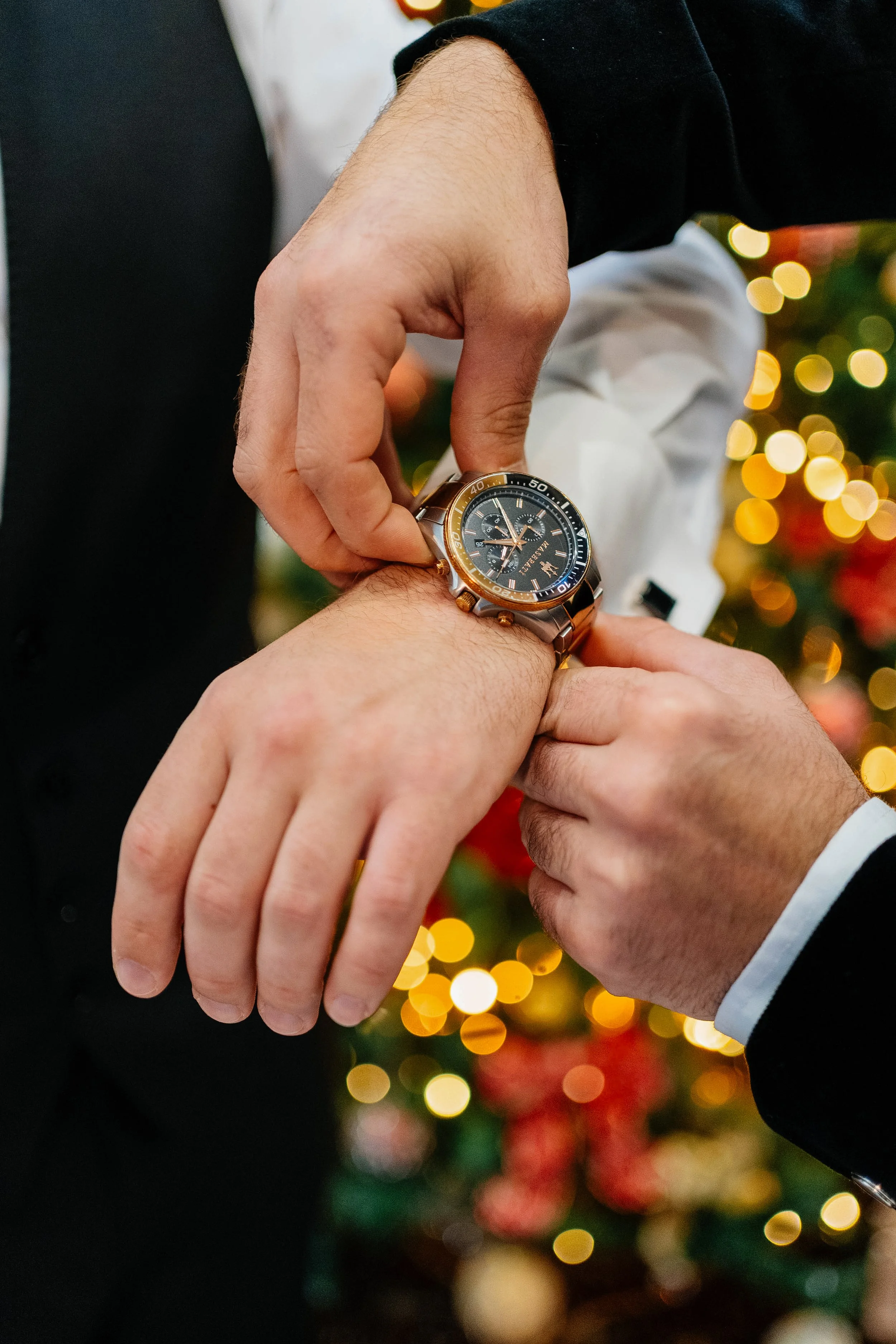 Two men dressed in formal attire exchanging rings during a wedding ceremony or celebration with blurred holiday lights in the background.
