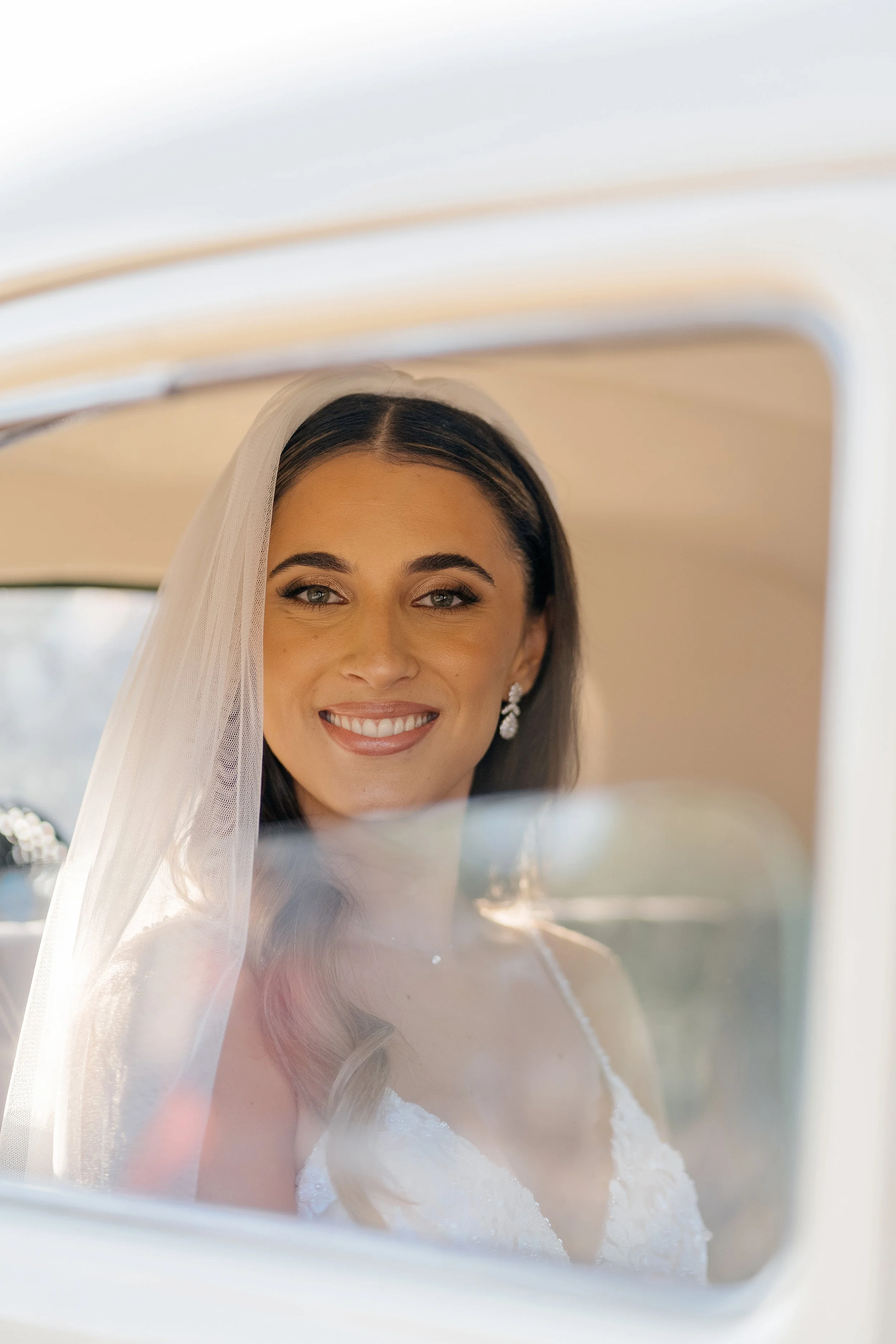 Close-up of a smiling bride with dark hair, wearing earrings and a veil, looking through a car window.