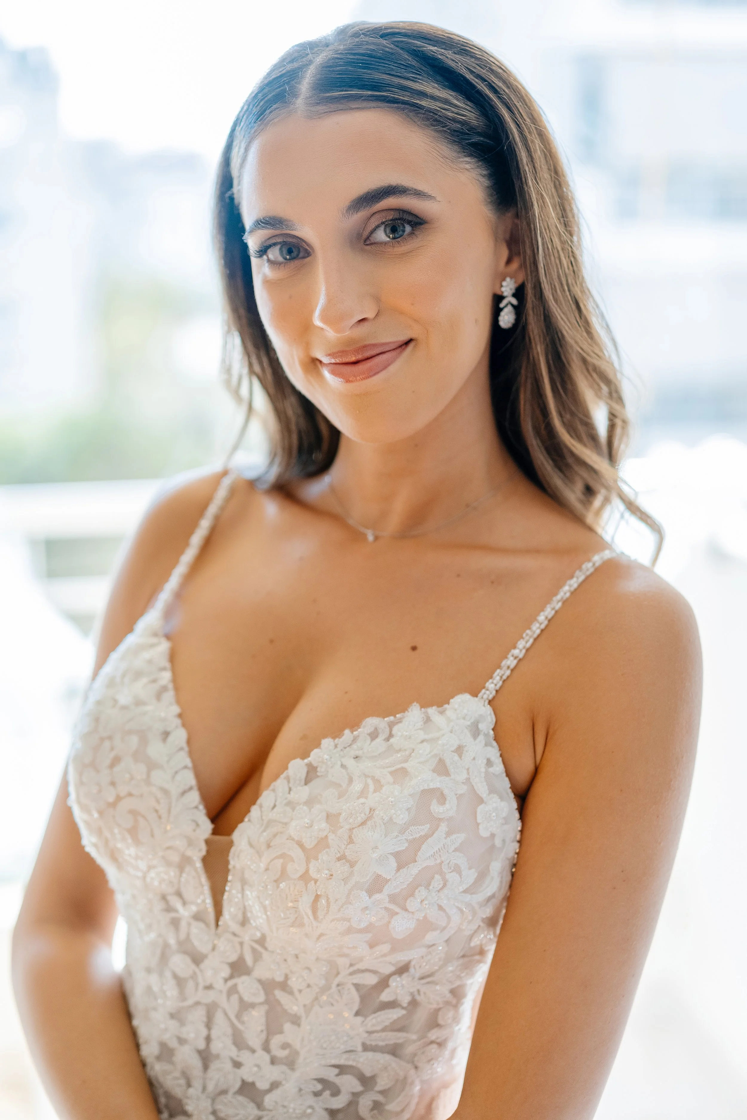 A woman wearing a white lace dress with floral embroidery, jewelry including earrings and a necklace, smiling with shoulder-length brown hair, in a well-lit room with large windows in the background.