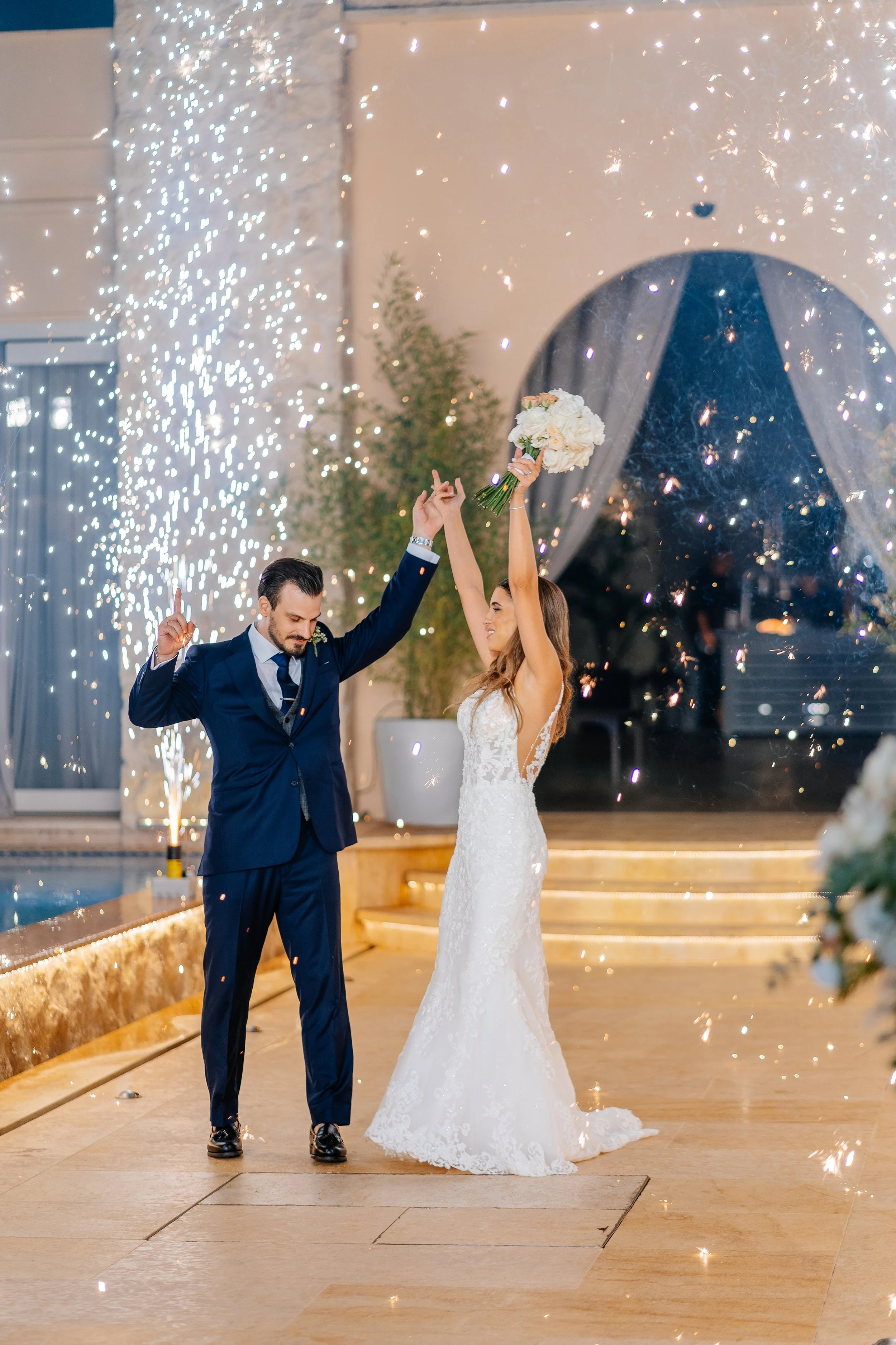 A bride and groom celebrating with a sparkler explosion. The bride is dressed in a white lace wedding gown, holding a bouquet, and the groom is in a dark blue suit and black shoes. They are indoors, standing on a light-colored tiled floor, with stair