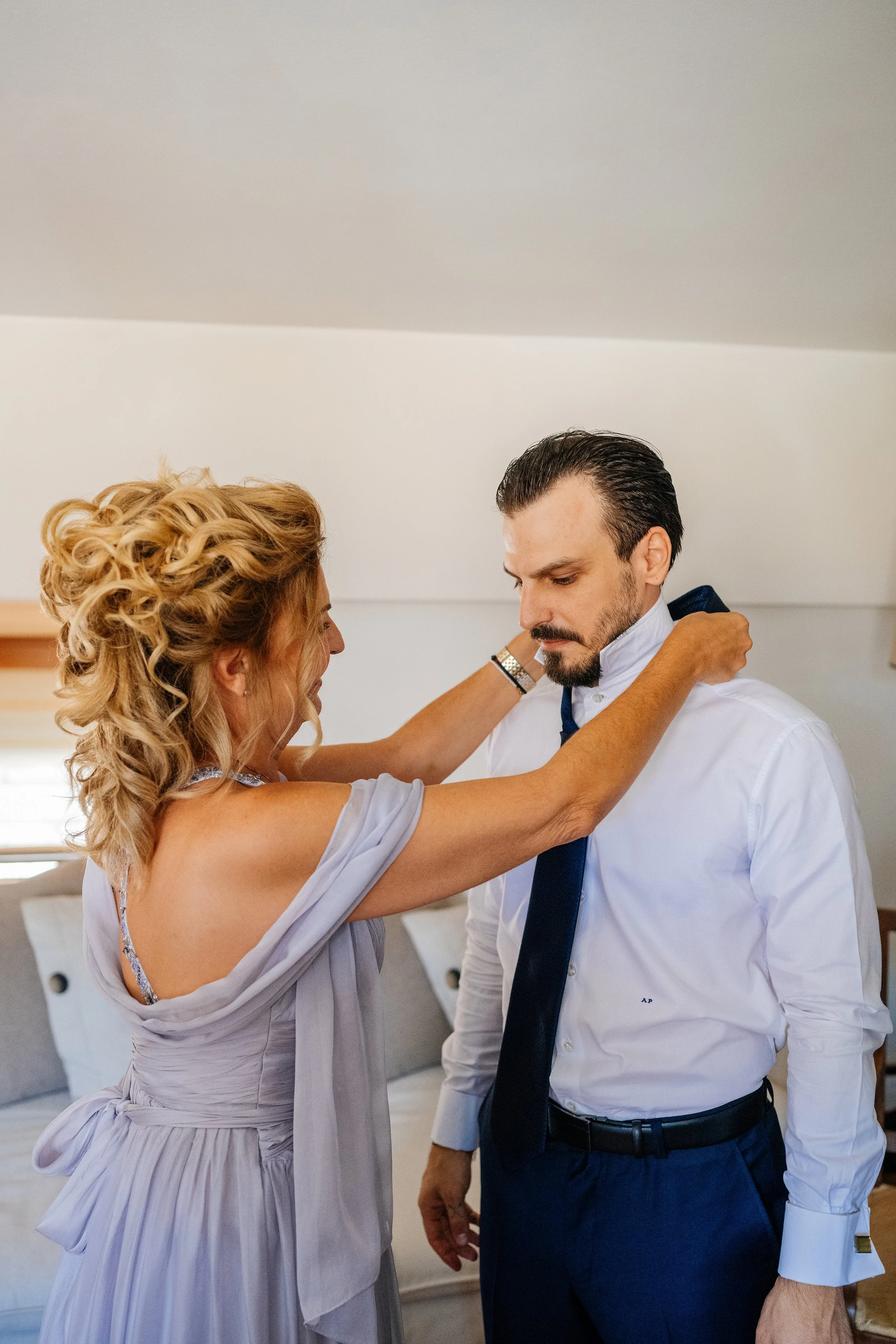 A woman with curly blonde hair helping a man adjust his tie in a bedroom, both dressed formally.