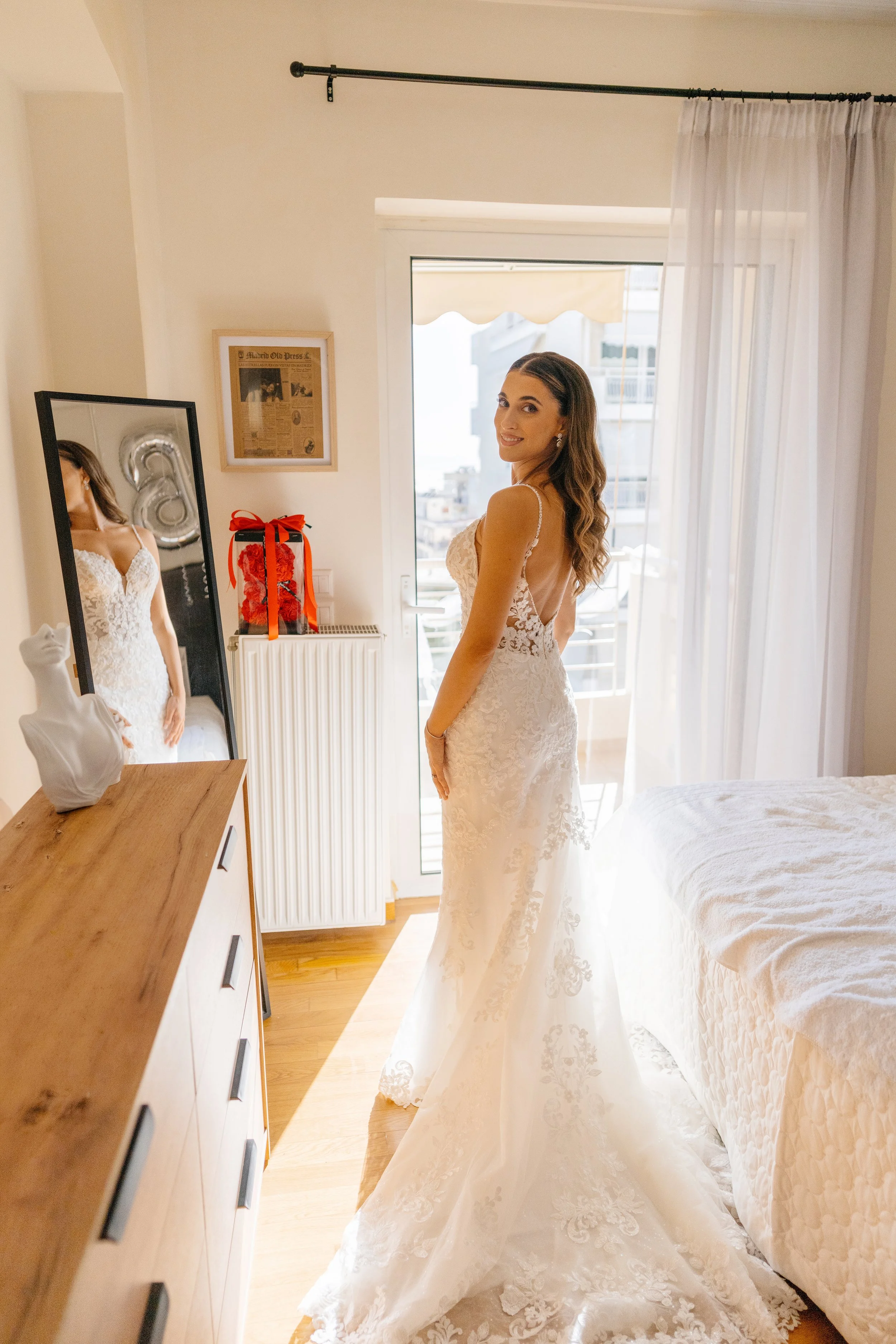 A woman in a wedding dress smiling in a well-lit bedroom near a window with curtains. There is a full-length mirror, a framed newspaper clipping, and a gift bag with a red ribbon on a dresser.