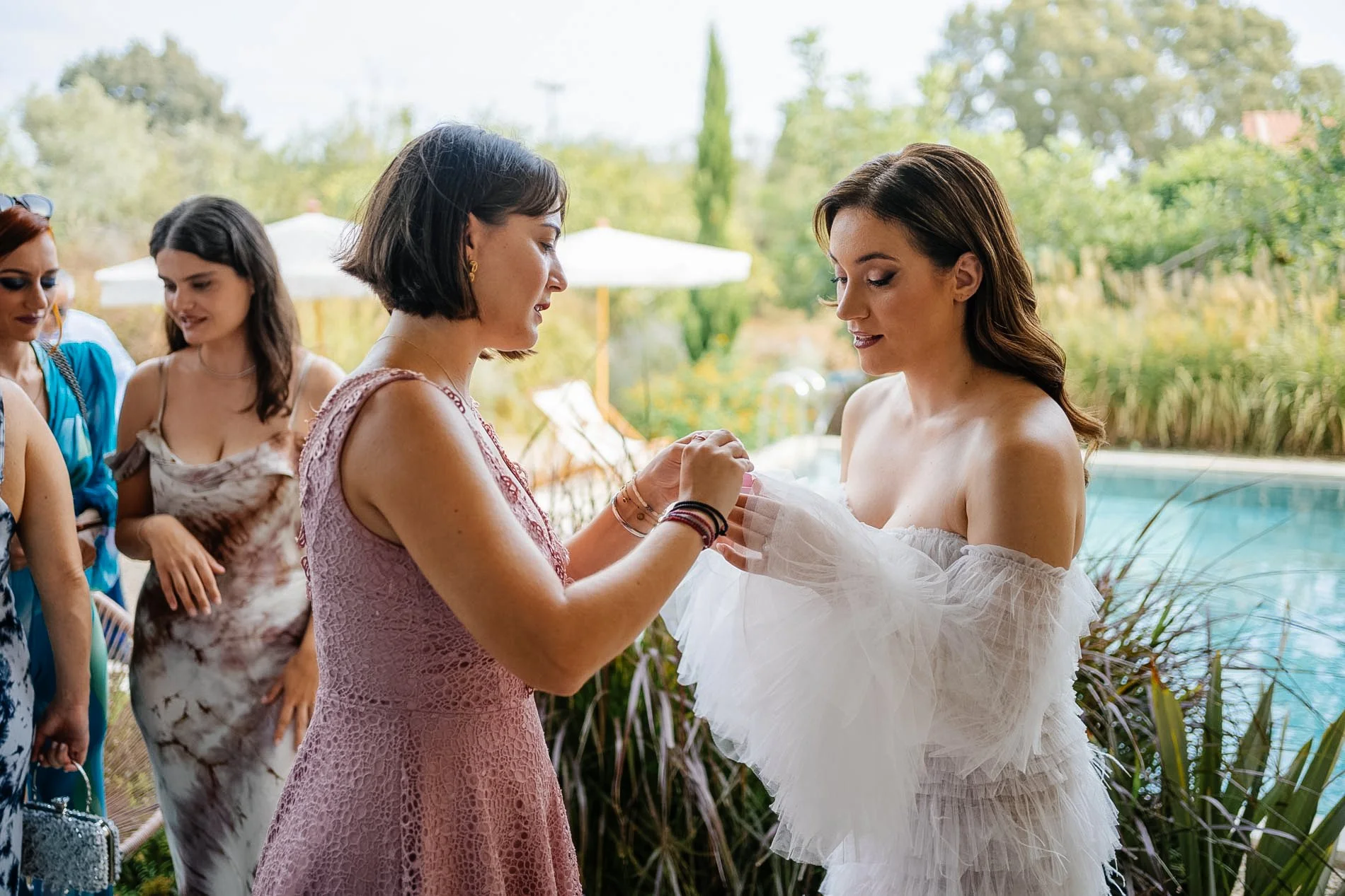 A woman in a white dress possibly at her wedding, receiving a bracelet from another woman. Several women are gathered around by the poolside, some smiling, with lush greenery in the background.