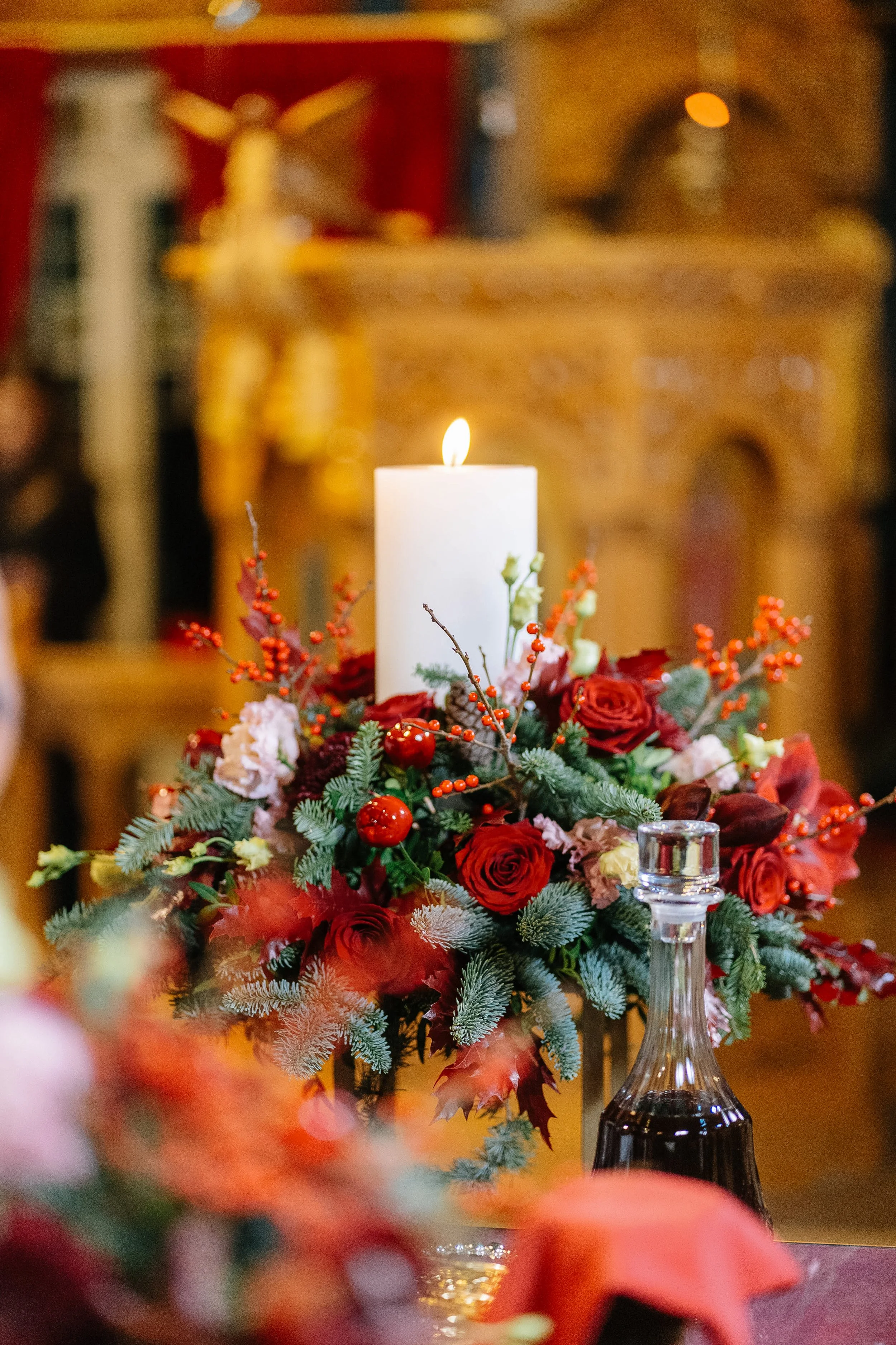 A holiday floral arrangement with a white candle on top, featuring red roses, green pine branches, red berries, and various flowers, set against a blurred background with warm lighting.