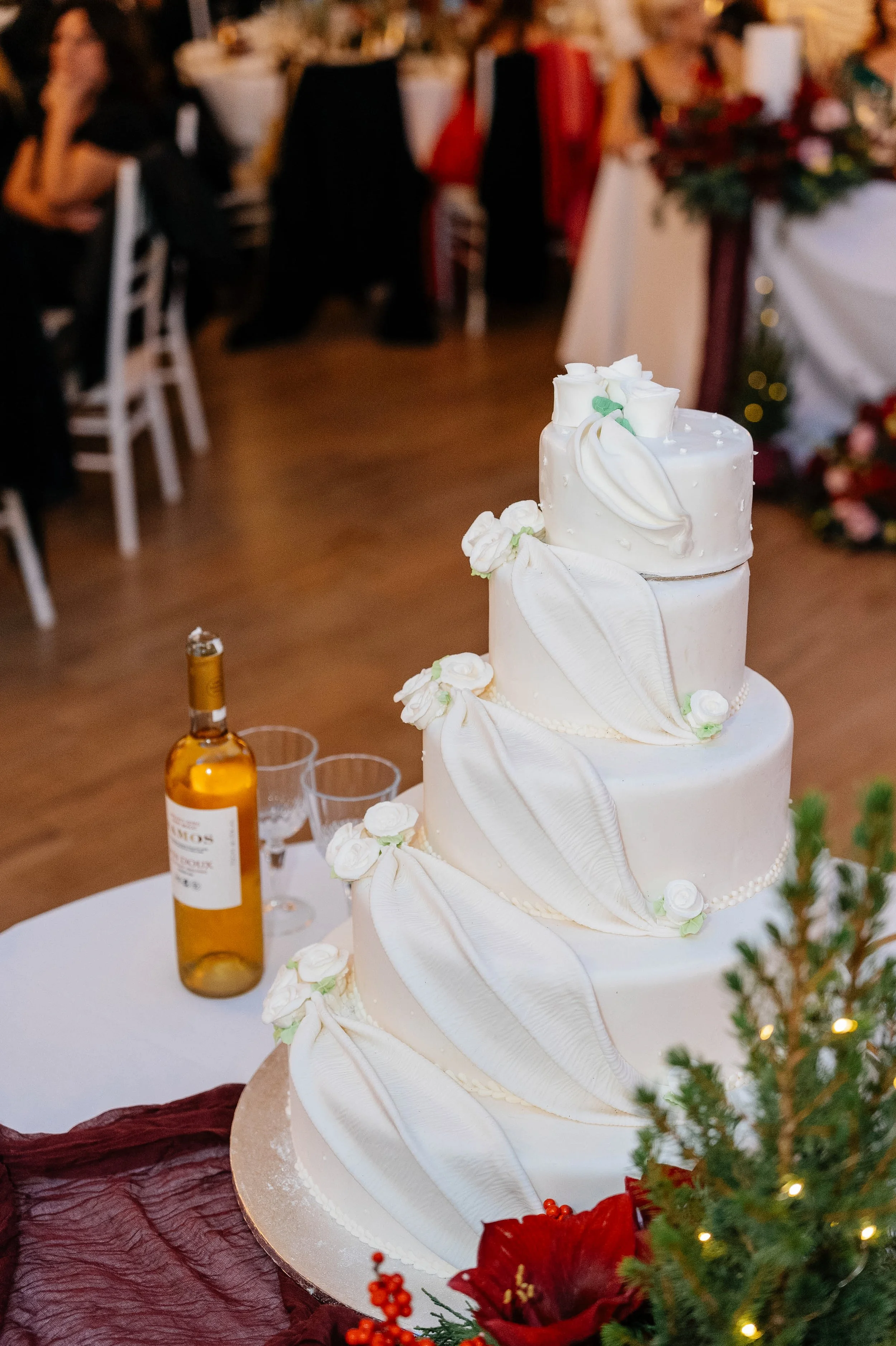A three-tier white wedding cake decorated with white flowers and draped fabric, set on a table with a bottle of alcohol and Christmas decorations, at a wedding reception.