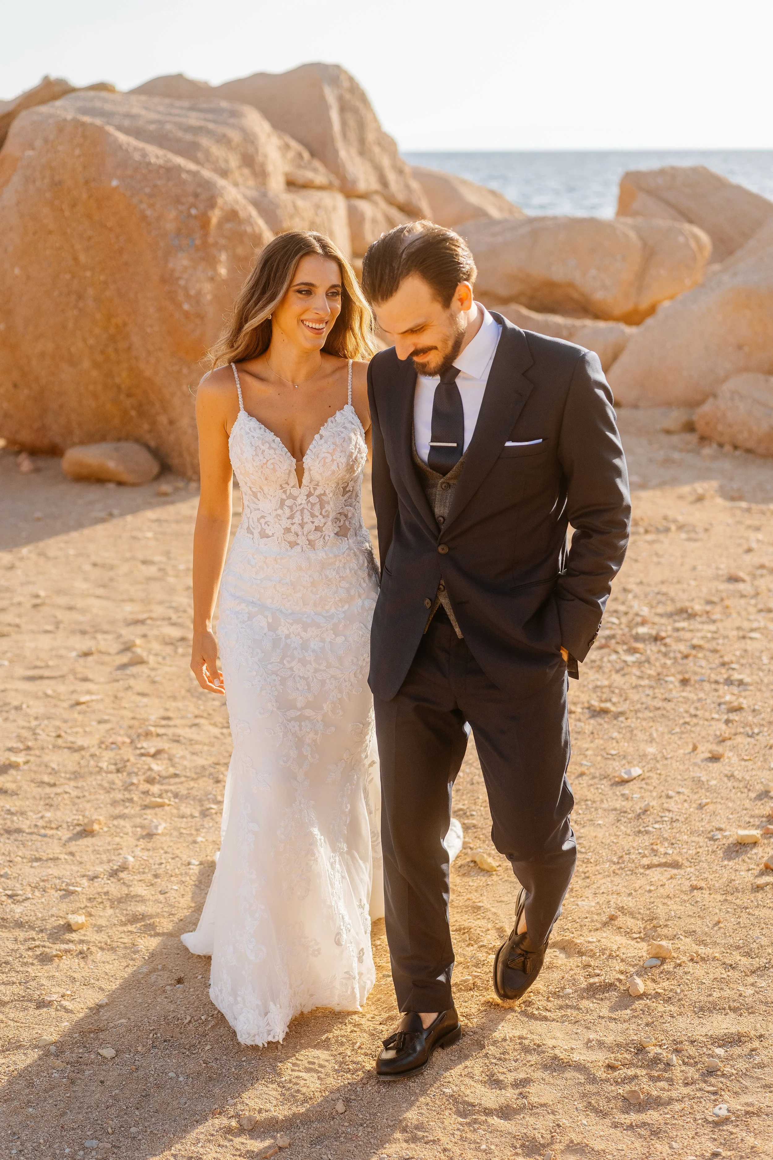 A bride in a white lace wedding gown and a groom in a dark suit with a white shirt and tie walking along a rocky beach with large sand-colored boulders and the ocean in the background during sunset.
