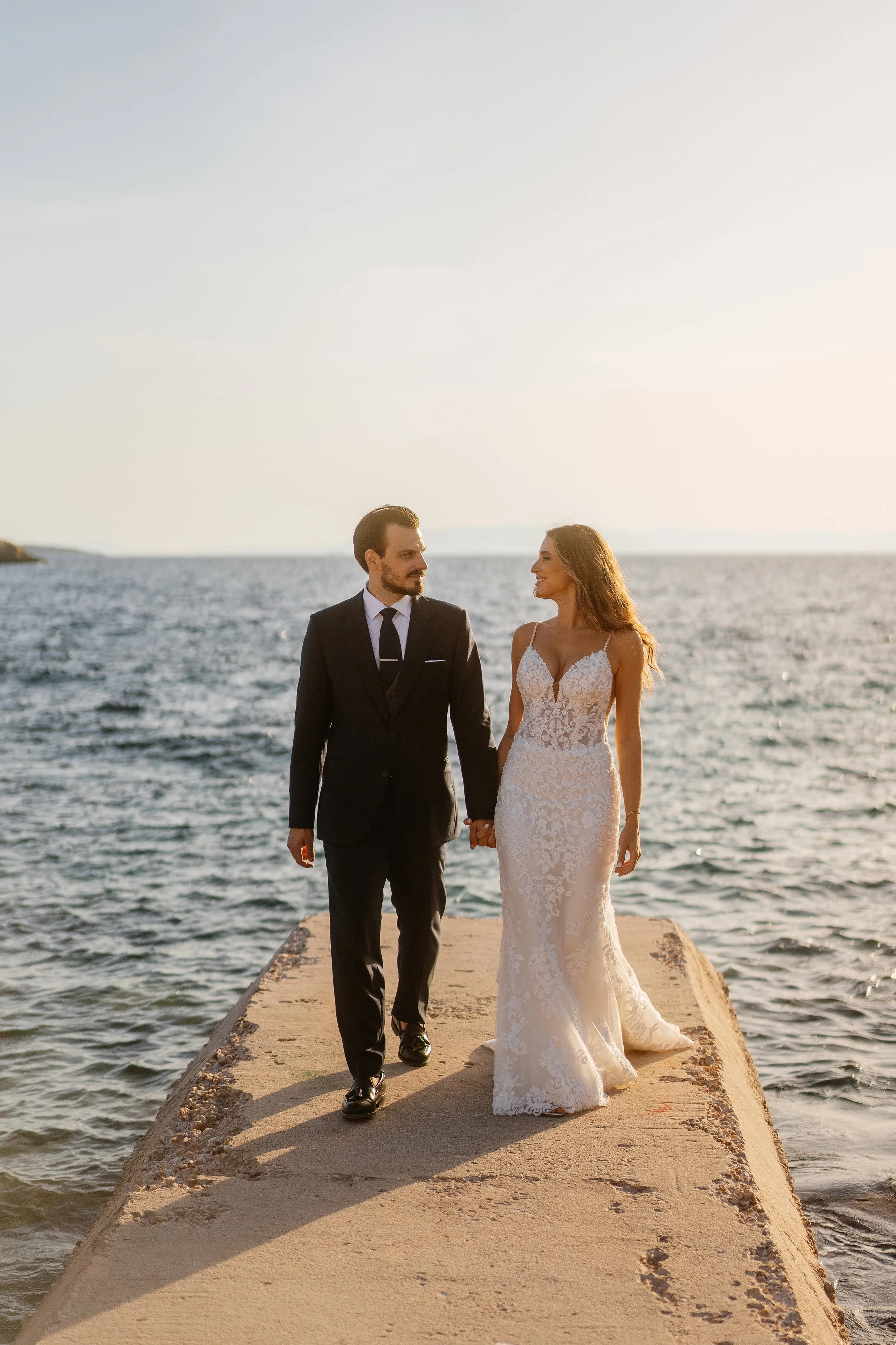 A bride and groom walking hand in hand on a concrete pier by the ocean during sunset. The groom wears a dark suit and tie, and the bride wears a white lace wedding dress. They are smiling and looking at each other.