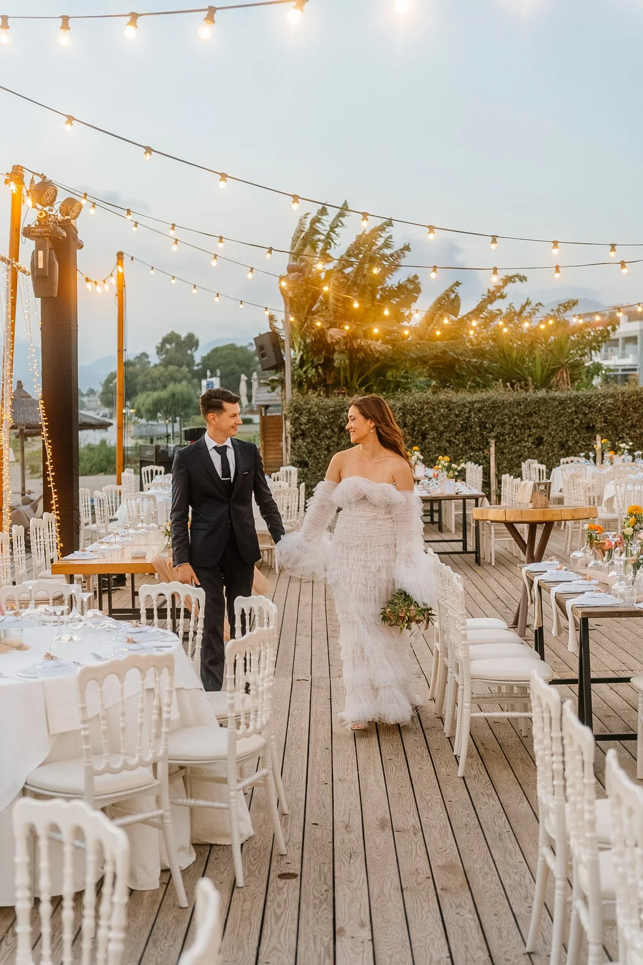 A bride and groom walk hand in hand through a decorated outdoor reception area during sunset, with string lights overhead and tables set for a wedding reception.