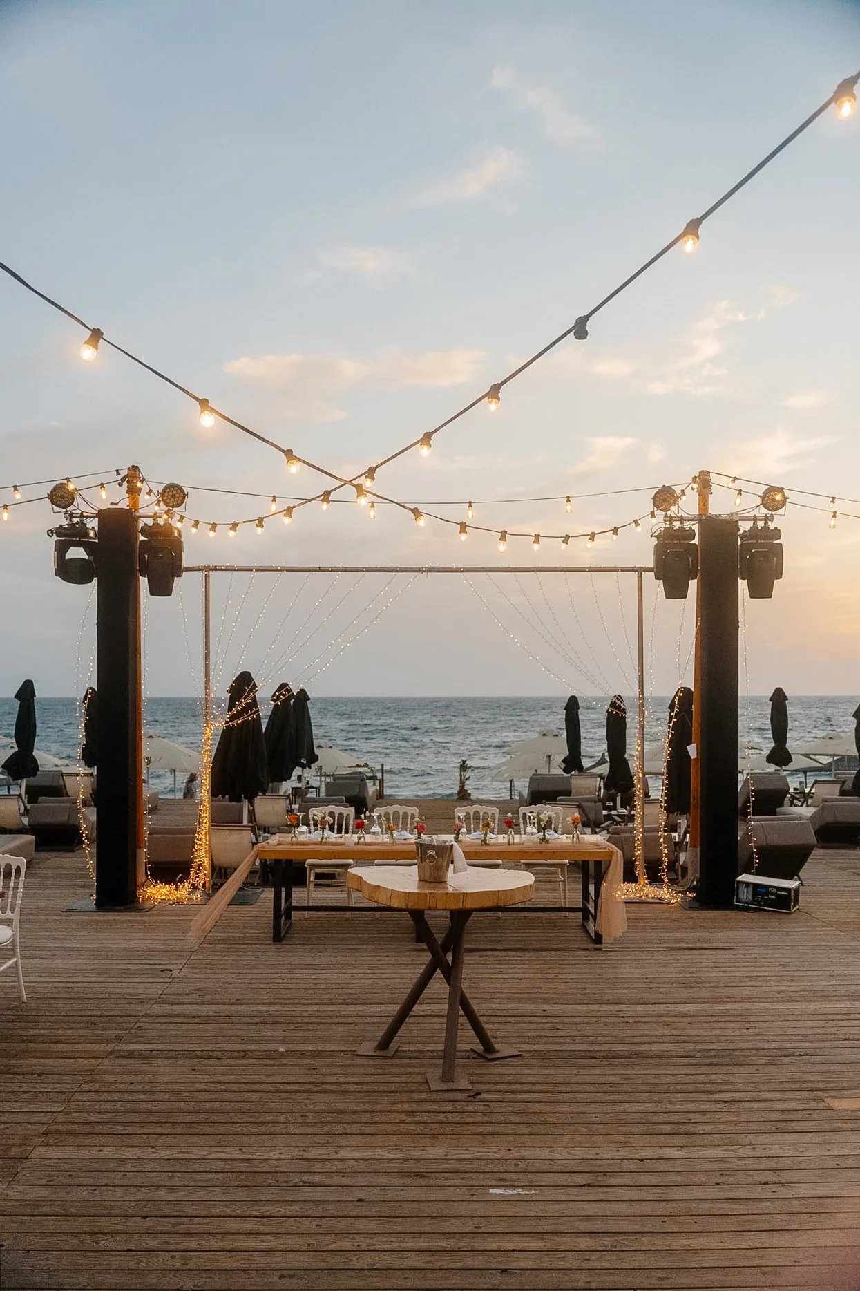 Beachside outdoor event setup with string lights, tables, chairs, and umbrellas, overlooking the ocean at sunset.