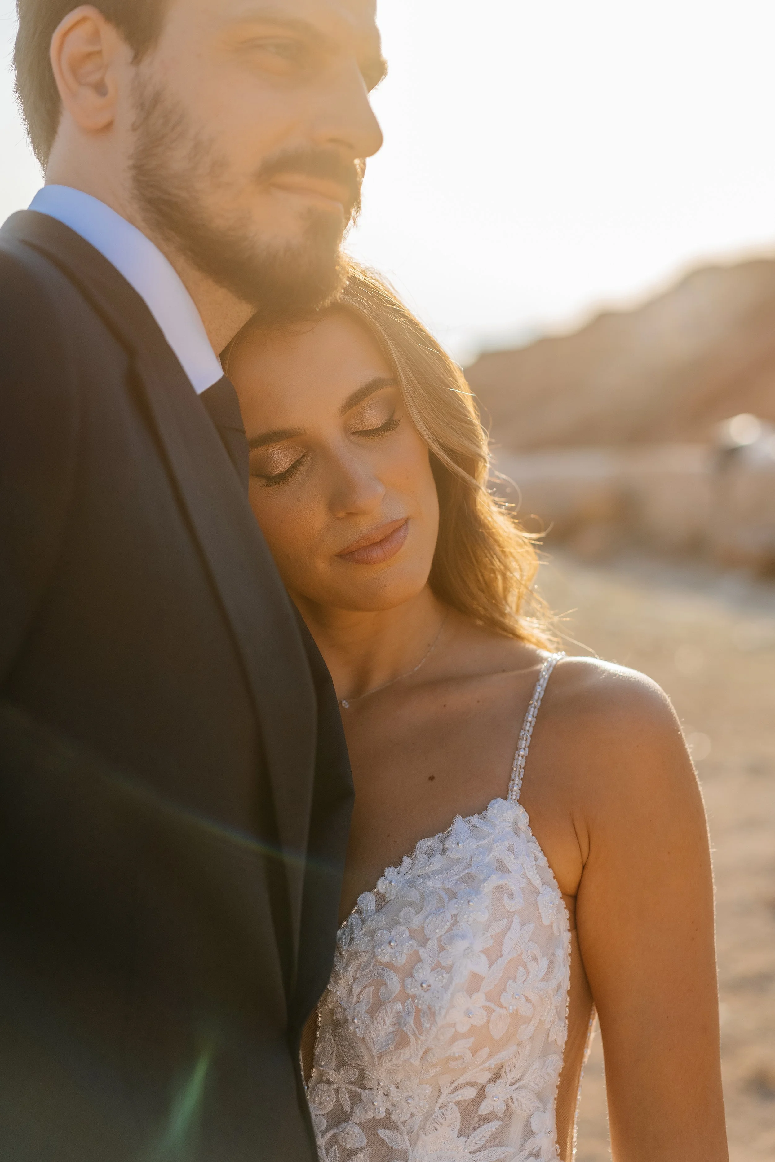 A man in a tuxedo and a woman in a white lace wedding dress embracing outdoors at sunset.