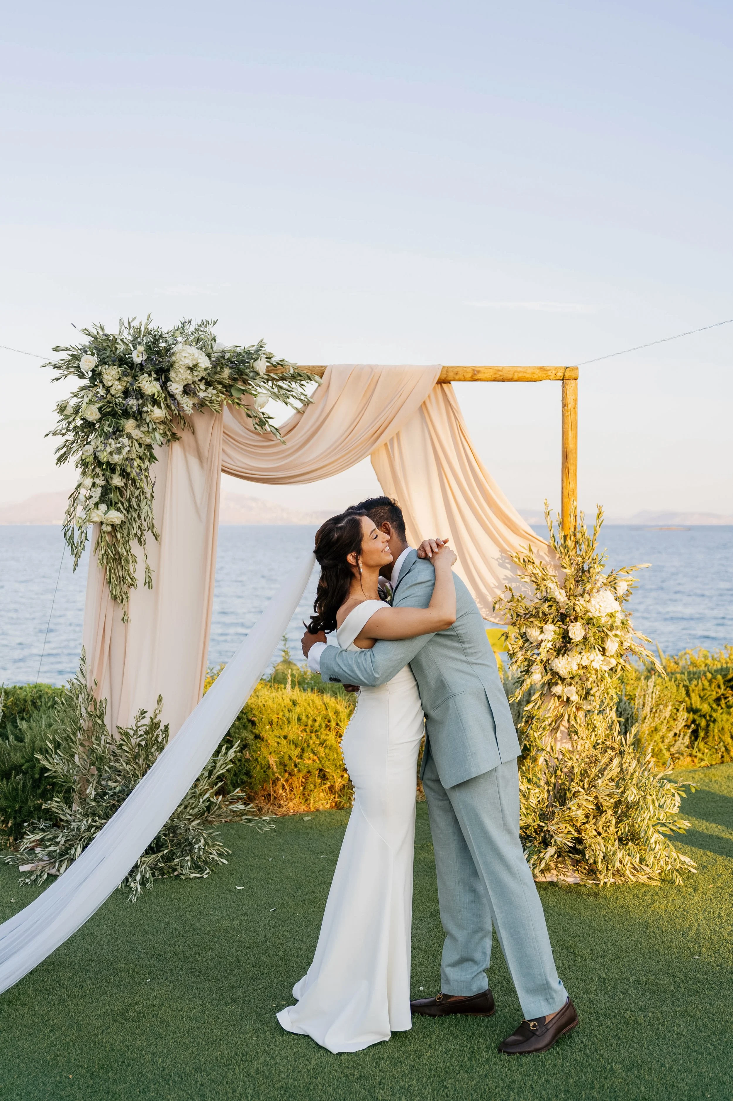A bride and groom embrace during their outdoor wedding ceremony at a scenic location by the water, with a floral decorated wooden arch.
