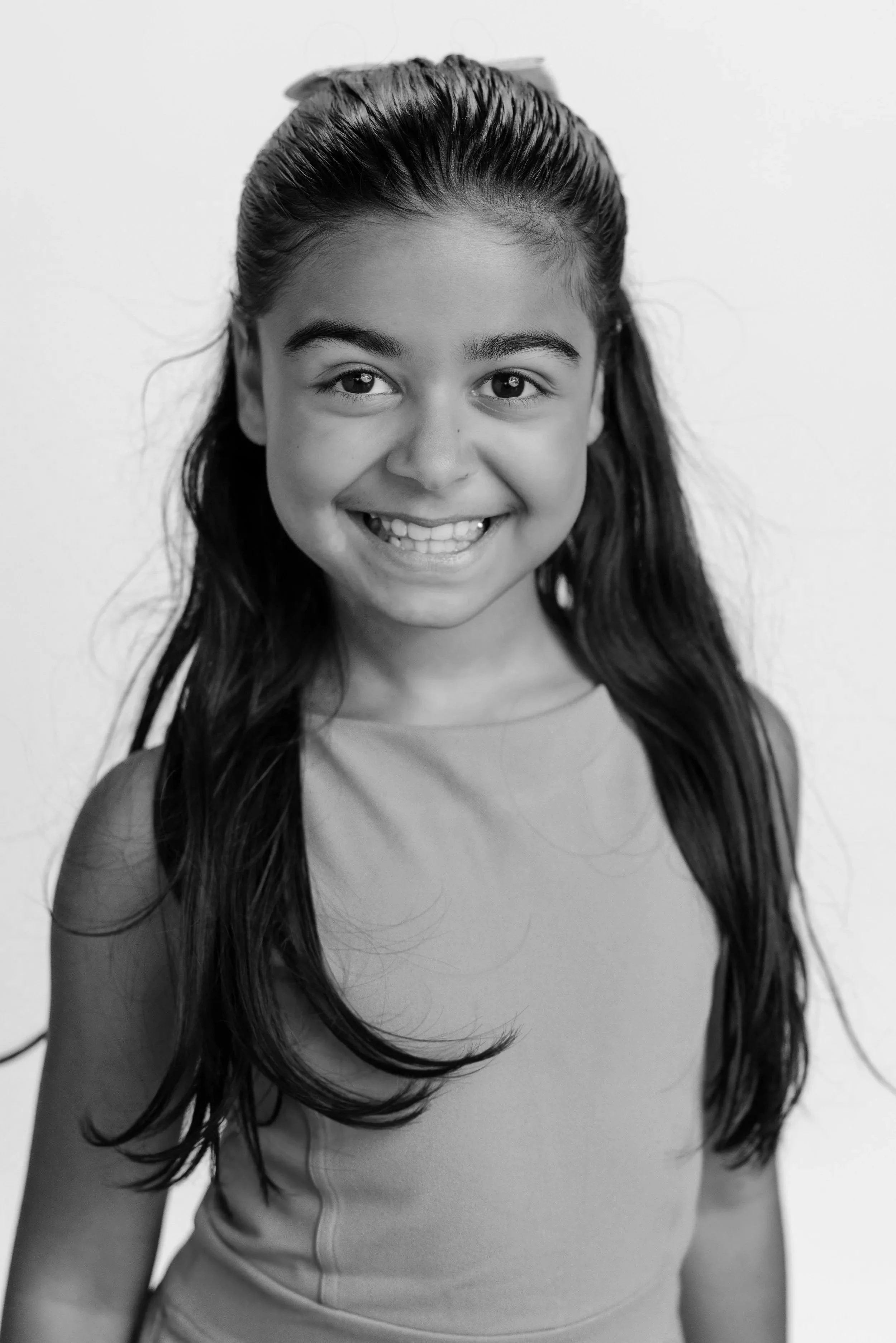 A young girl with long dark hair smiling at the camera, wearing a sleeveless top, in black and white.