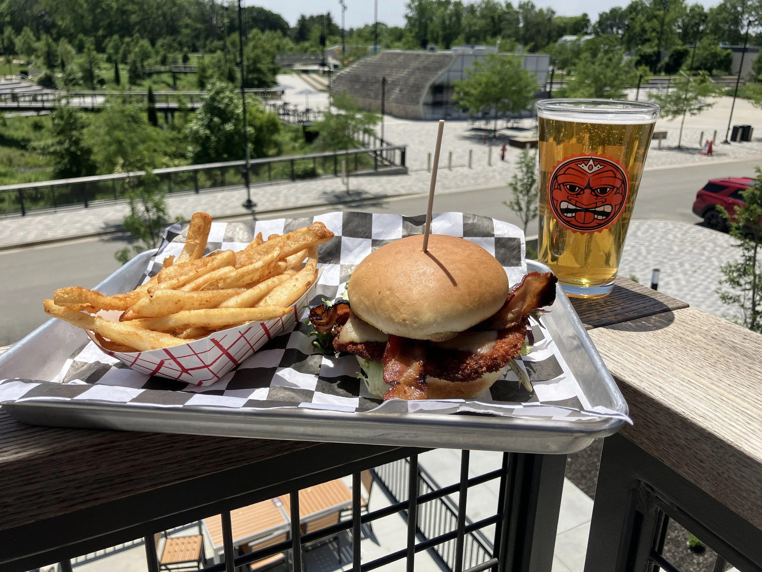 Tray with Stacked House tenderloin sandwich and fries with a Sun King Premium Light Lager in a pint glass sitting on table on the balcony of sun king Westfield overlooking the Grand Junction Park.