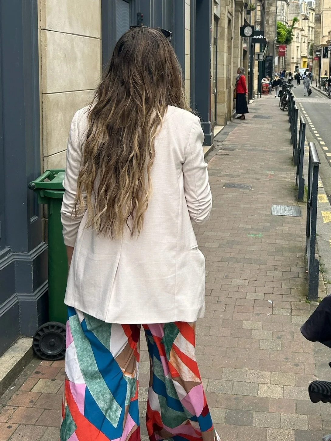 Une femme avec de longs cheveux bruns portant un blazer beige et un pantalon coloré à motifs marche dans une rue de ville.
