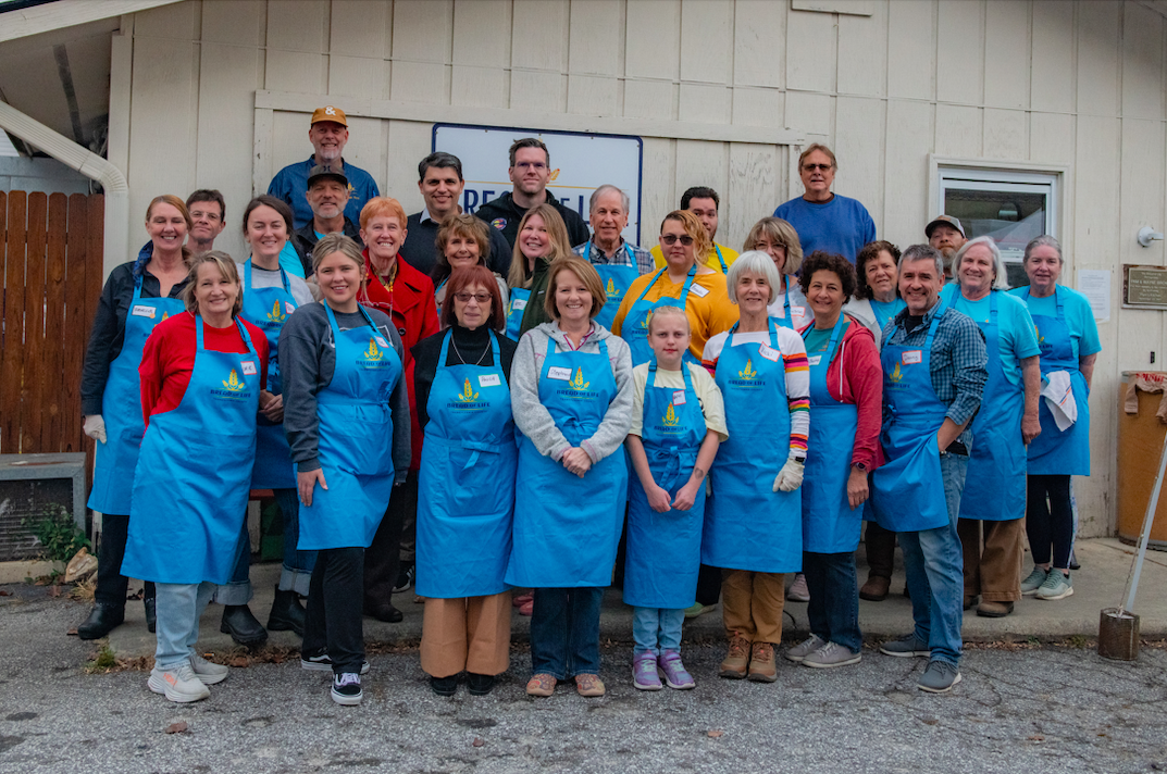 A group of people gathered for a fall-themed event inside a room with green walls, decorated with pumpkins and autumn leaves on the table. Some people are wearing aprons and name tags, posing for the photo.