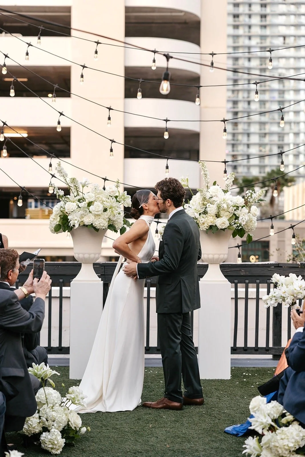 A bride and groom kiss during their wedding ceremony on an outdoor terrace decorated with large classic timeless floral arrangements and string lights, with urban buildings in the background.